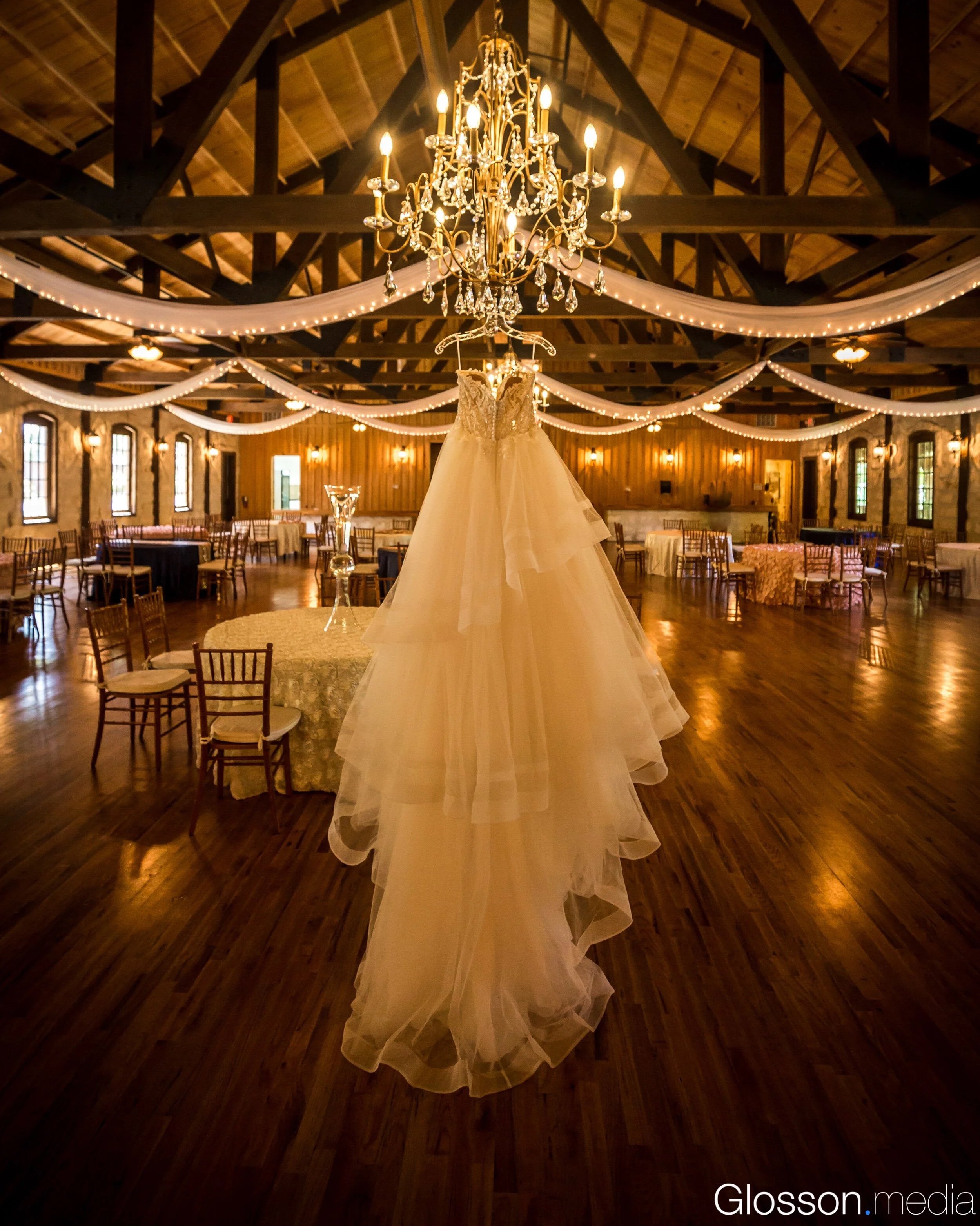 Wedding dress hanging from a chandelier in a decorated venue with wooden walls and ceiling, string lights, and tables set for a celebration.