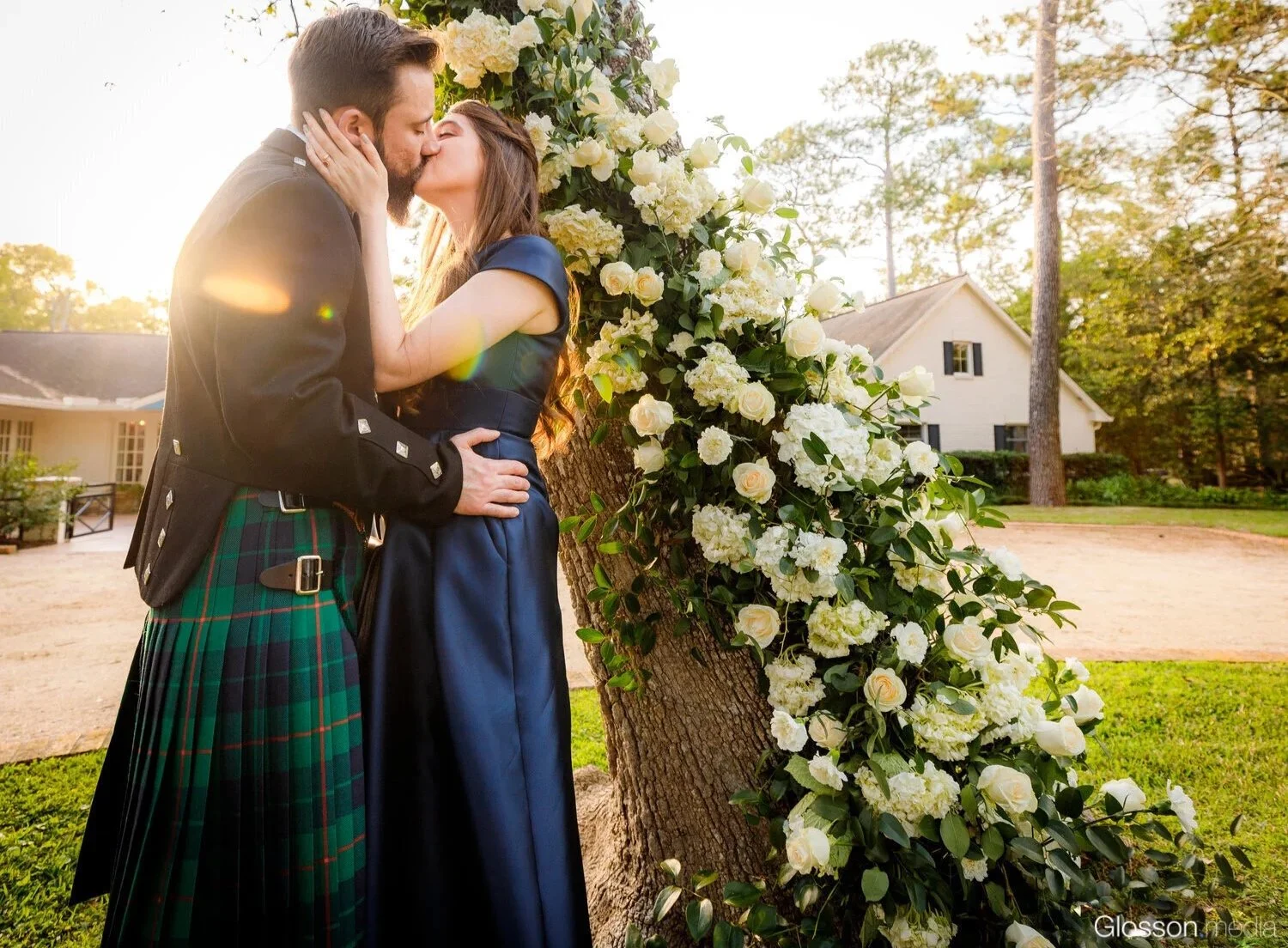 A couple sharing a kiss during their wedding outdoors at sunset, with a large flower arrangement on a tree behind them, and a small white house visible in the background.