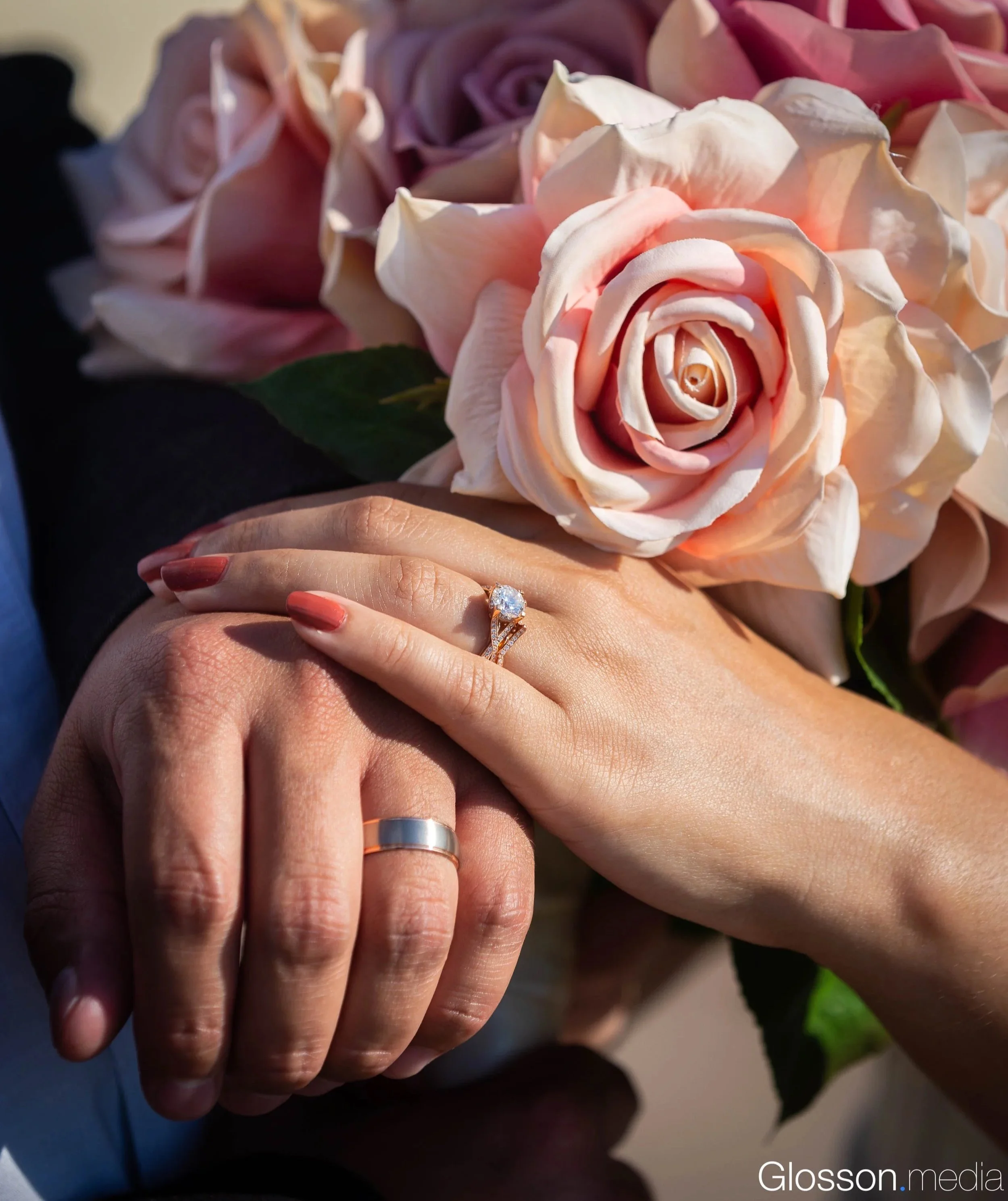 Close-up of two hands wearing wedding rings, with a large bouquet of pink and purple roses in the background.
