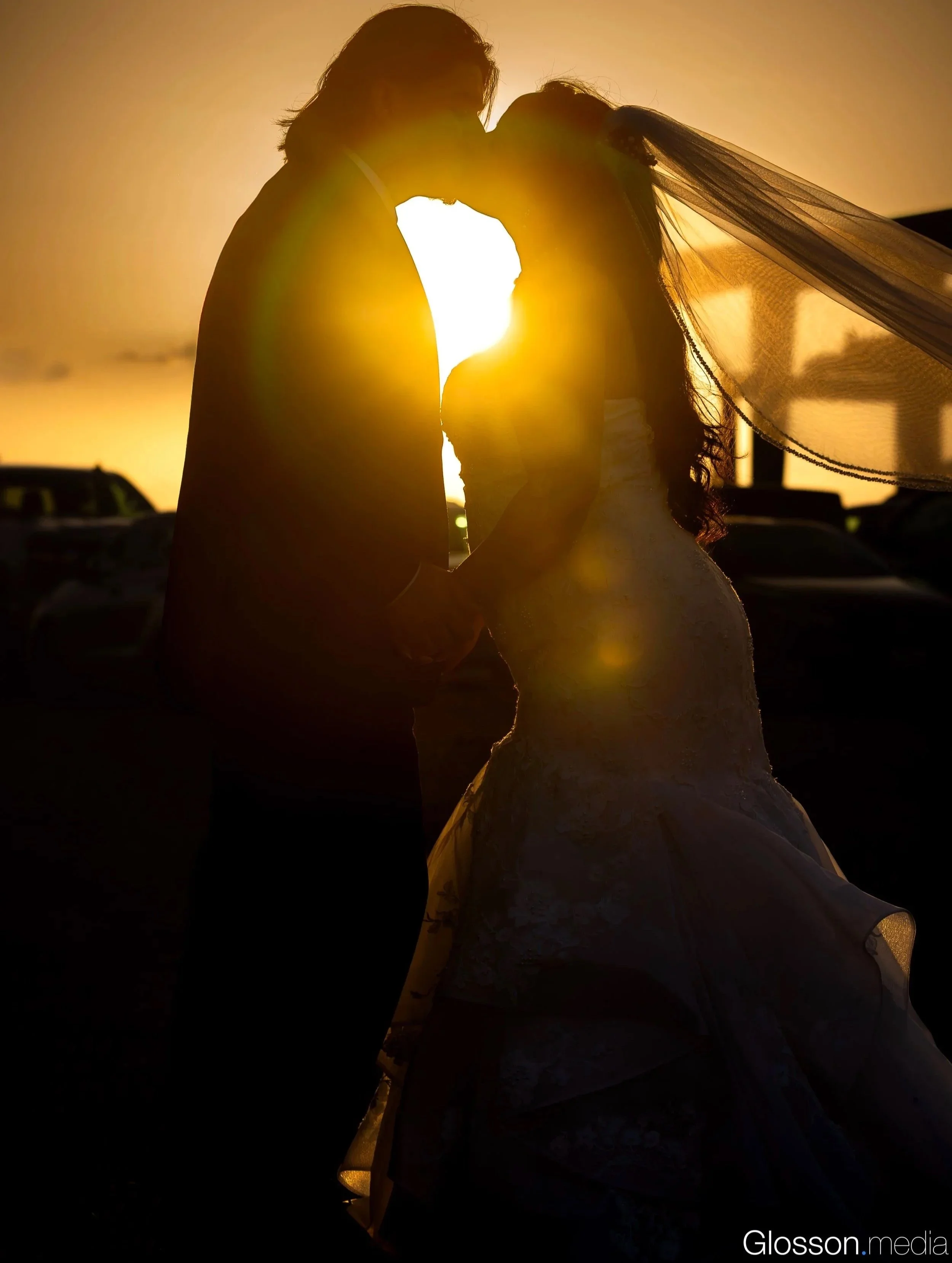 A silhouette of a couple kissing during sunset, with the bride wearing a veil and wedding gown, and the groom in dark suit, holding hands.