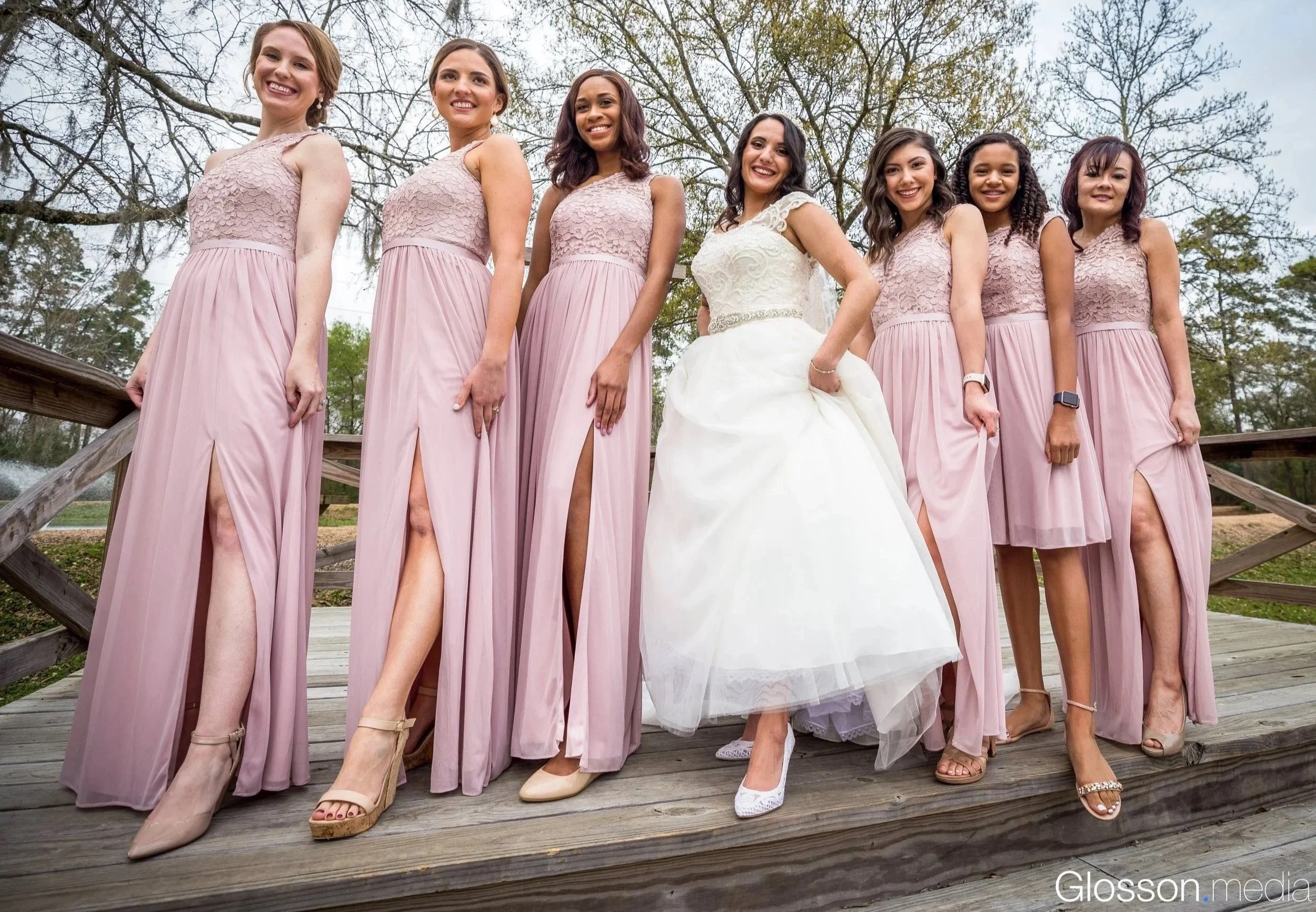 A bride in a white wedding gown surrounded by six bridesmaids in pink dresses, standing on a wooden bridge outdoors with trees and a cloudy sky in the background.