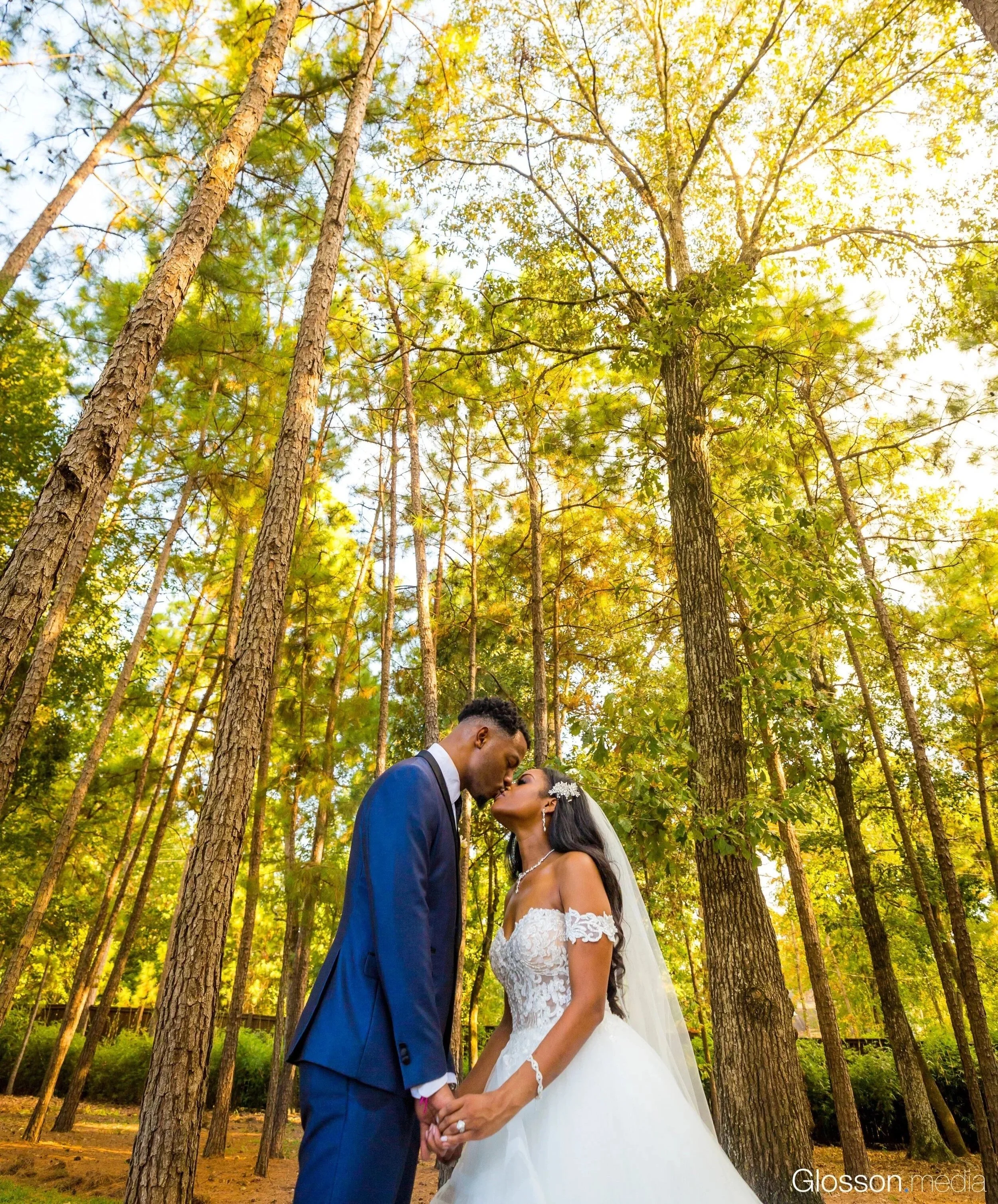 A bride and groom holding hands and leaning in for a kiss in a forest clearing with tall trees and sunlight filtering through.