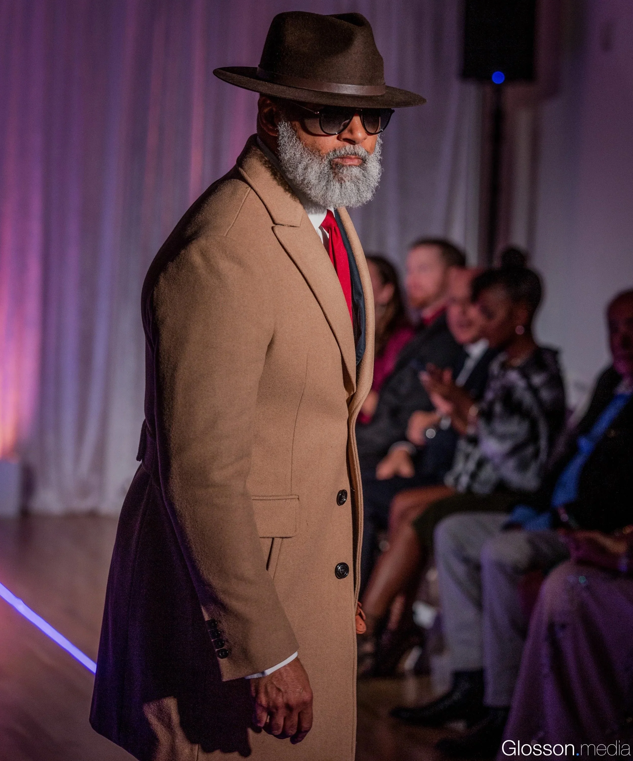 An elderly man with a gray beard dressed in a beige coat, white shirt, and red tie, wearing dark sunglasses and a wide-brimmed brown hat, standing in a dimly lit room with purple and pink lighting.