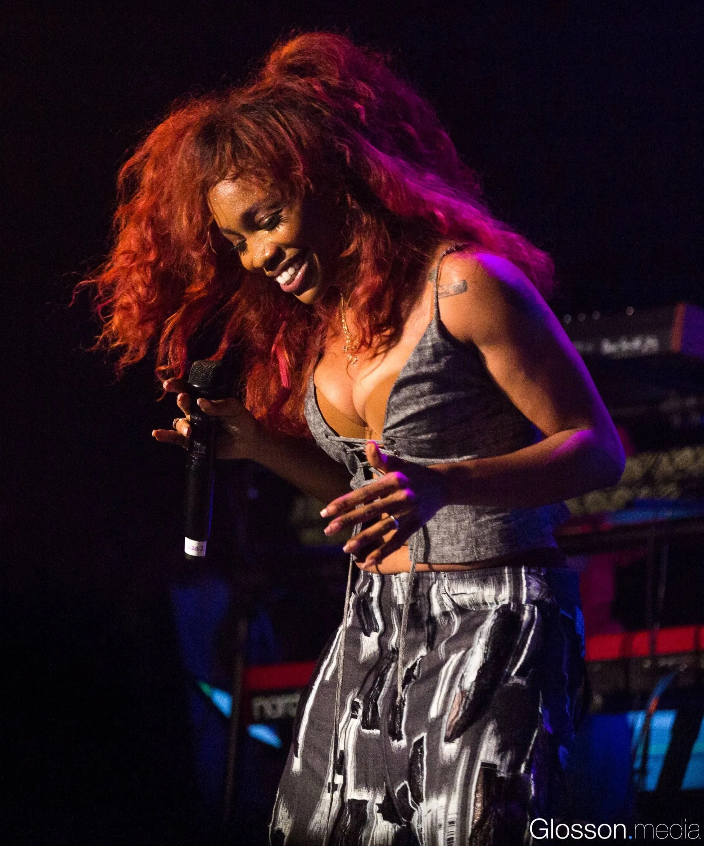 A woman with red curly hair holding a microphone, smiling, on stage with dark background and musical equipment in the background.