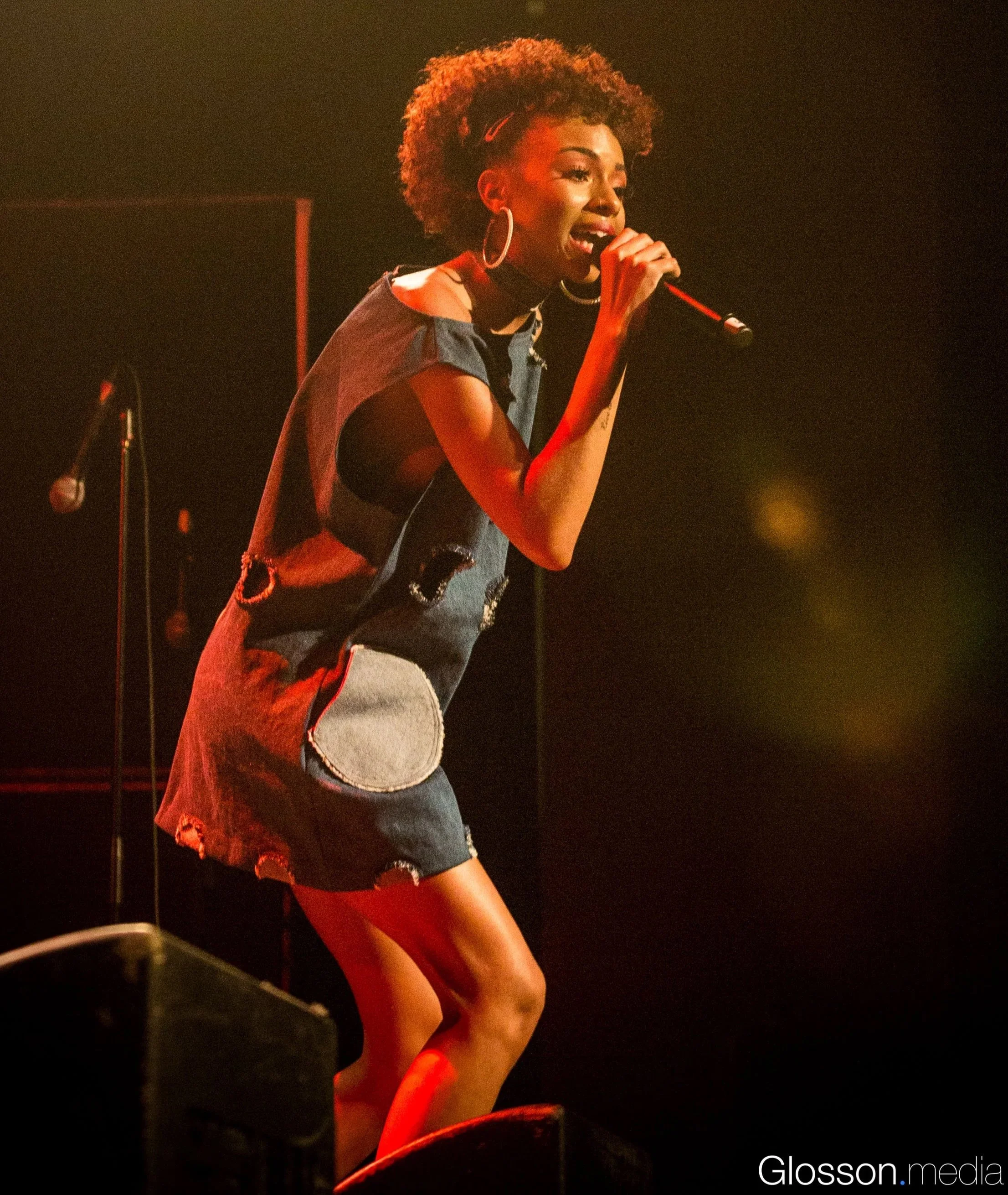 A woman singing on stage with a microphone, wearing a distressed denim dress with patches, large hoop earrings, and a natural hairstyle.