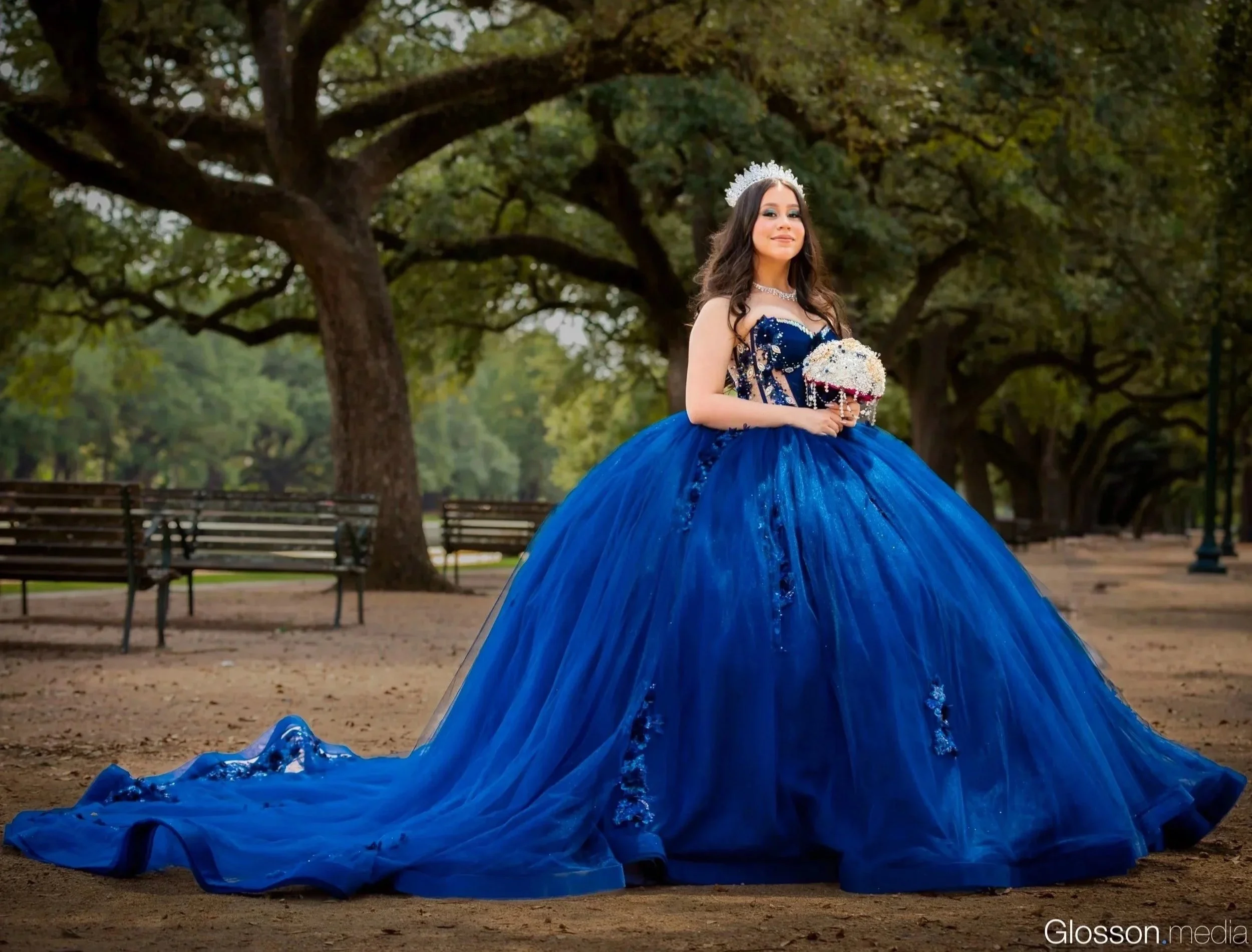 Girl in an elegant blue ball gown holding a bouquet, standing in a park with large trees and benches.