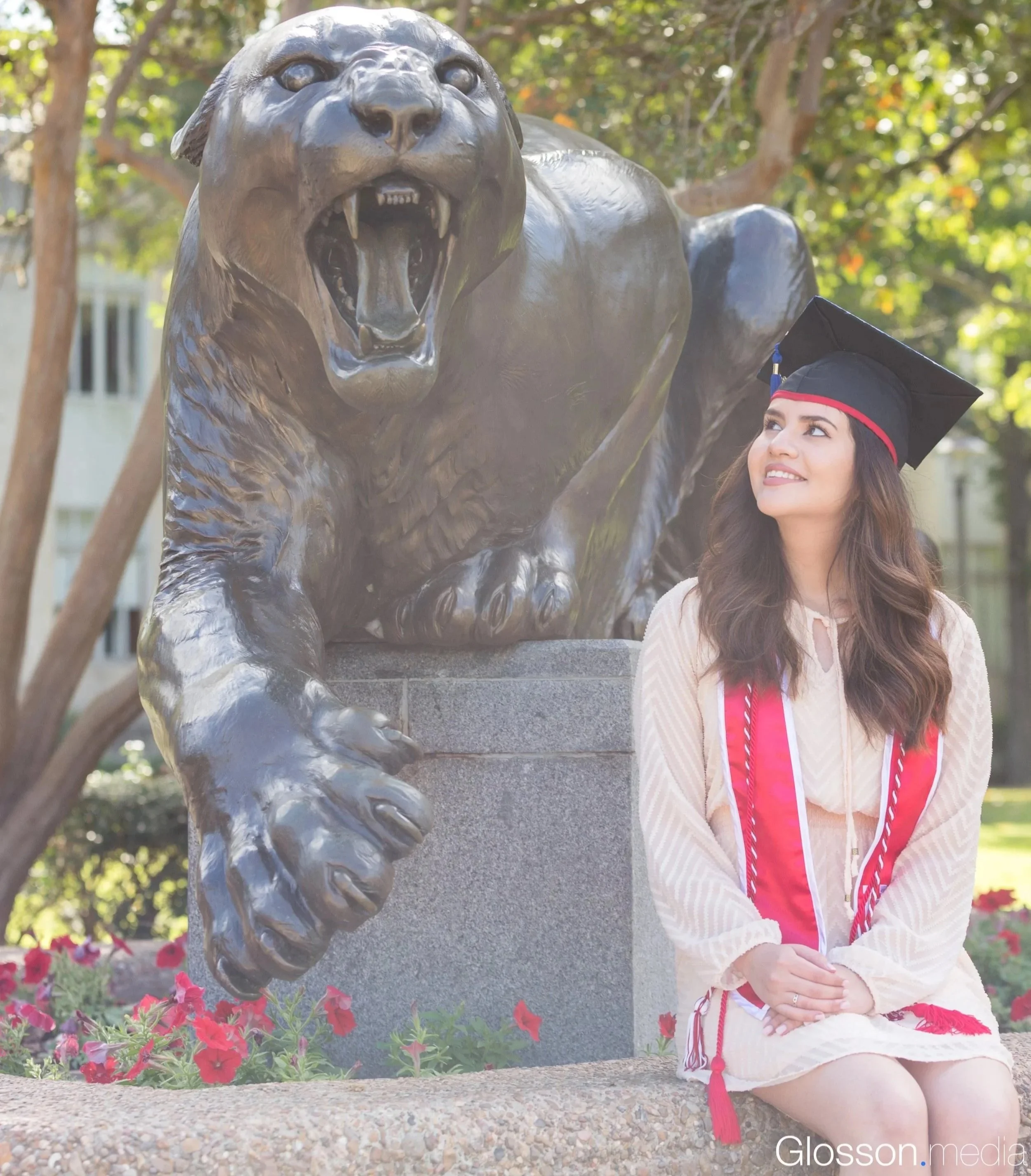 A young woman in graduation attire, including a cap and gown, sitting outdoors next to a large bronze lion statue. The woman is smiling and looking up at the lion. The background has trees and a building.