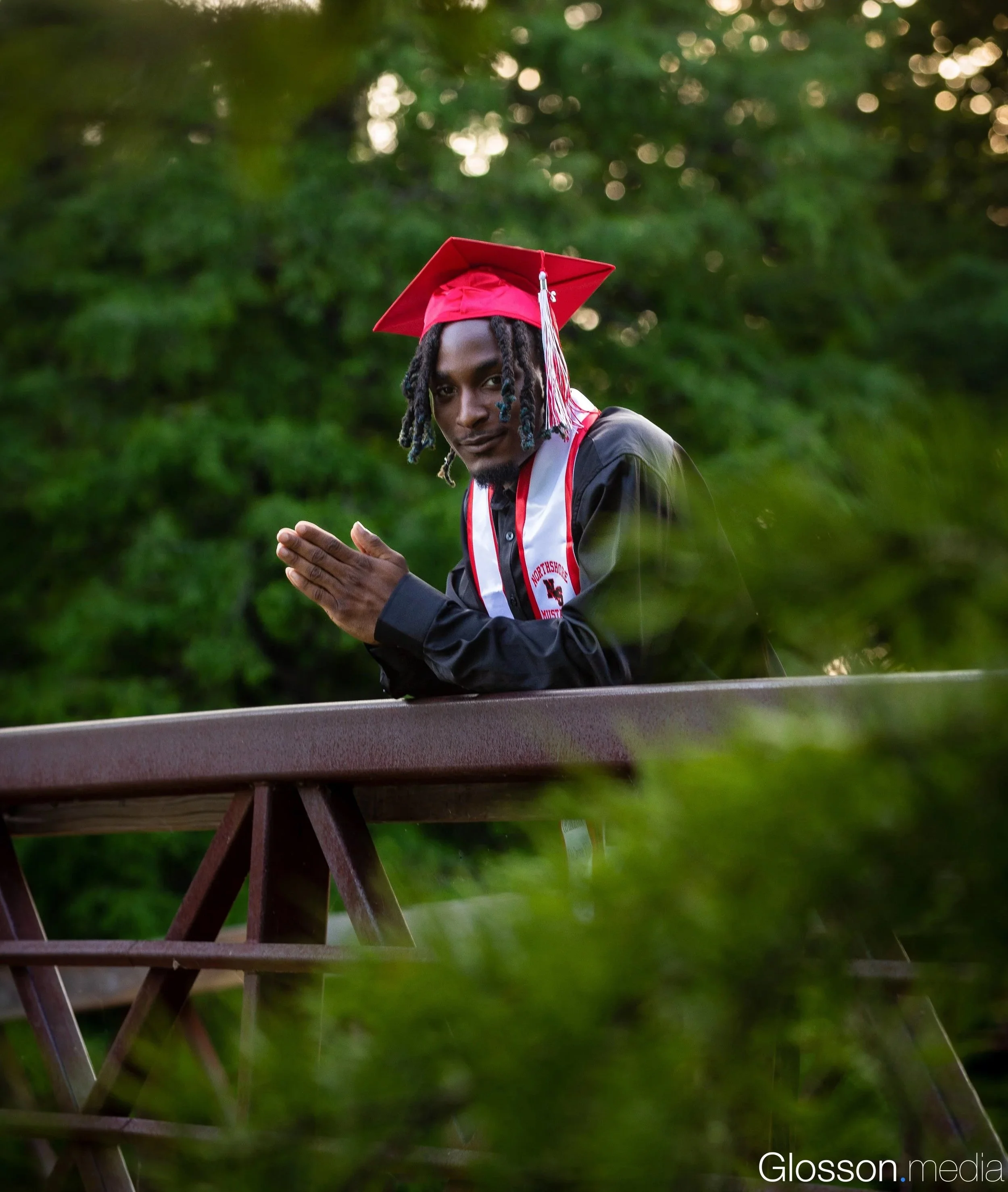 A young man in a black graduation gown and red cap with a tassel stands outdoors at sunset, leaning on a wooden railing with greenery in the background, hands clasped together in a gesture of gratitude or prayer.