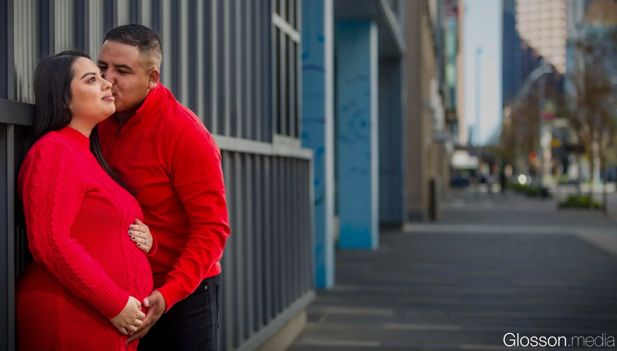 A couple in red clothing standing close to each other outdoors, with the man kissing the woman's forehead, leaning against a metal fence in an urban setting.