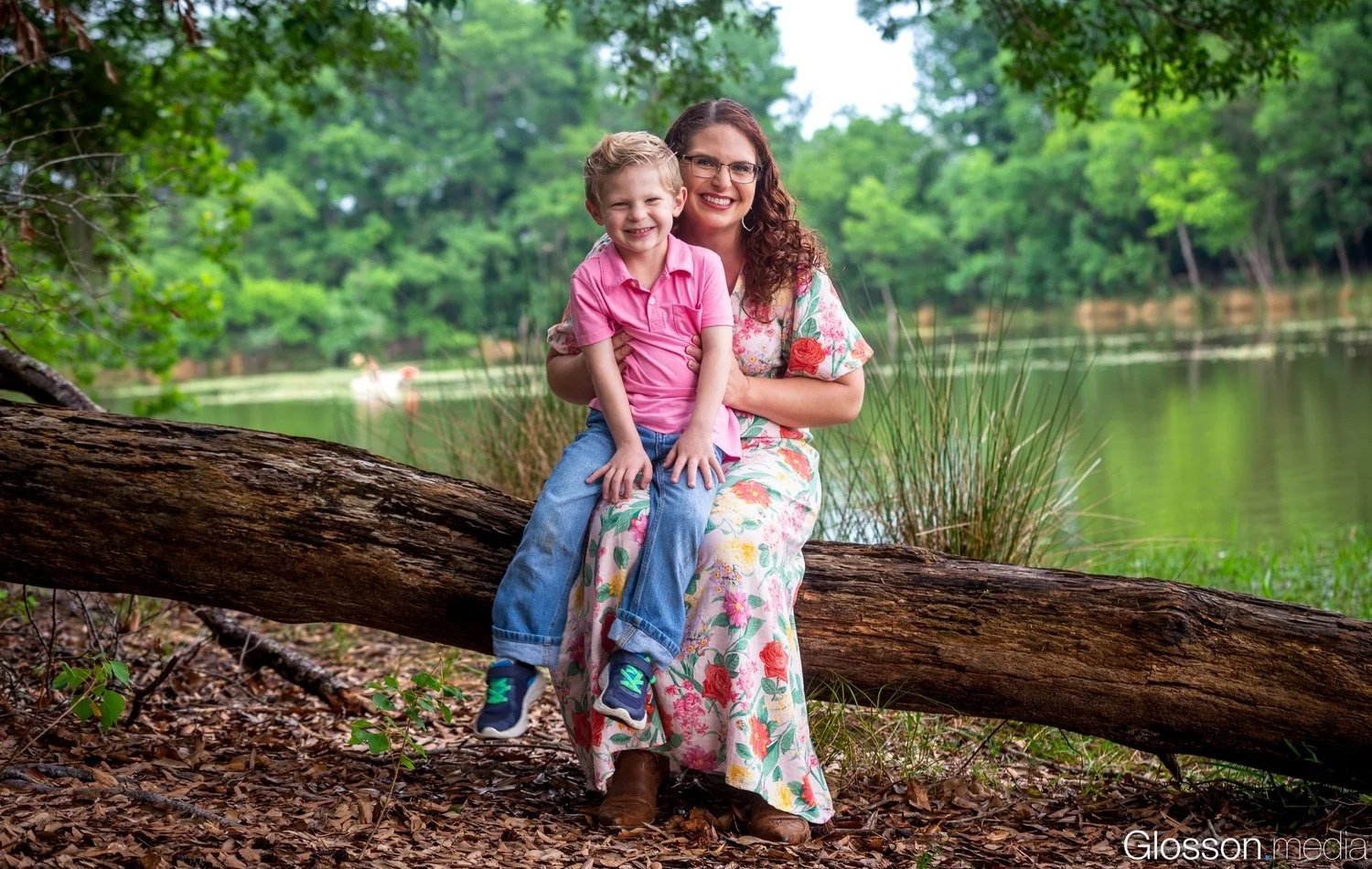 A woman and a young boy sitting on a fallen log by a lake in a wooded area, smiling at the camera.