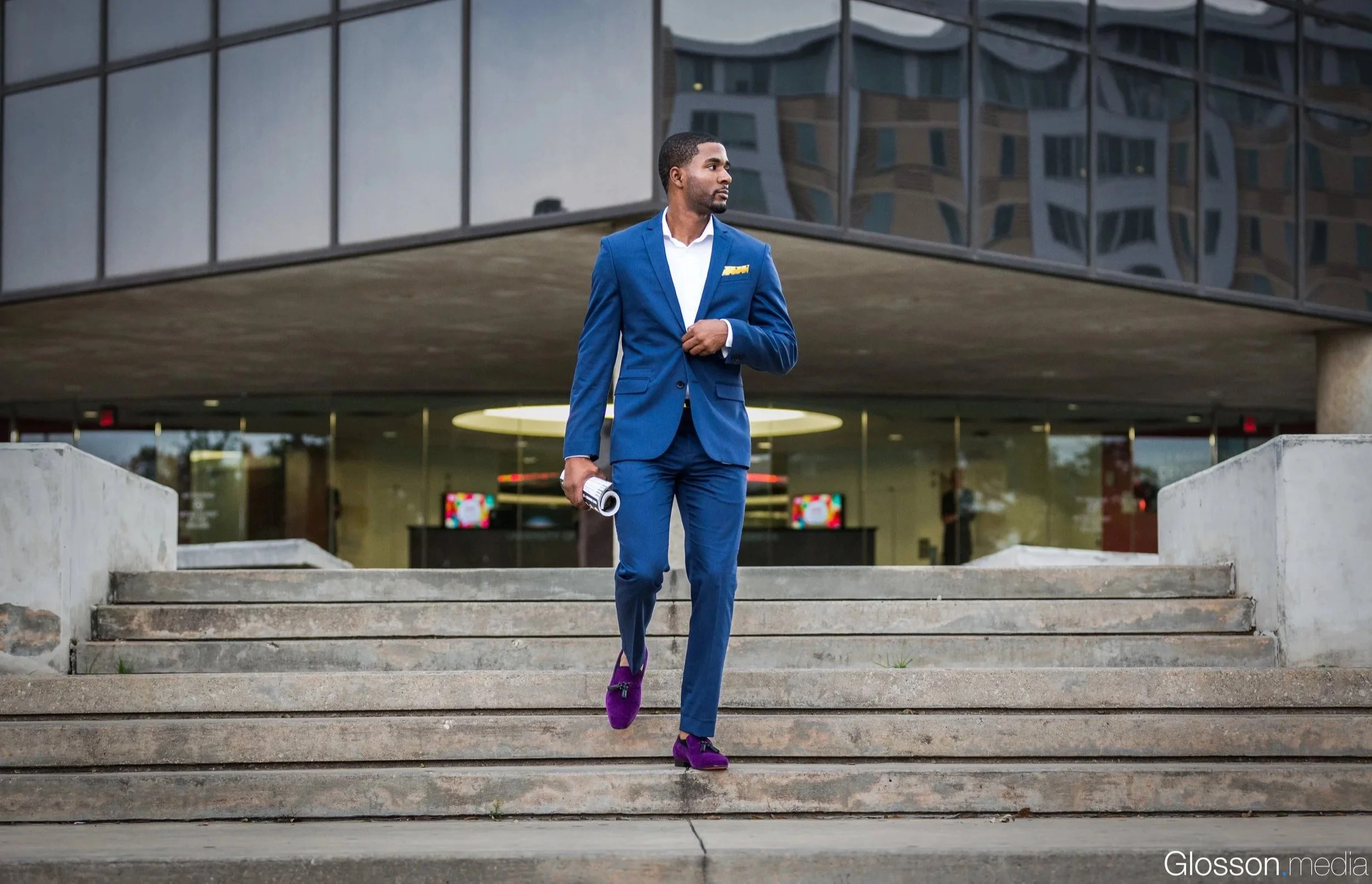 A man in a sharp blue suit with a yellow pocket square walks down concrete stairs outside a modern building with reflective glass windows. He is holding a rolled-up document in his right hand and wearing purple shoes.