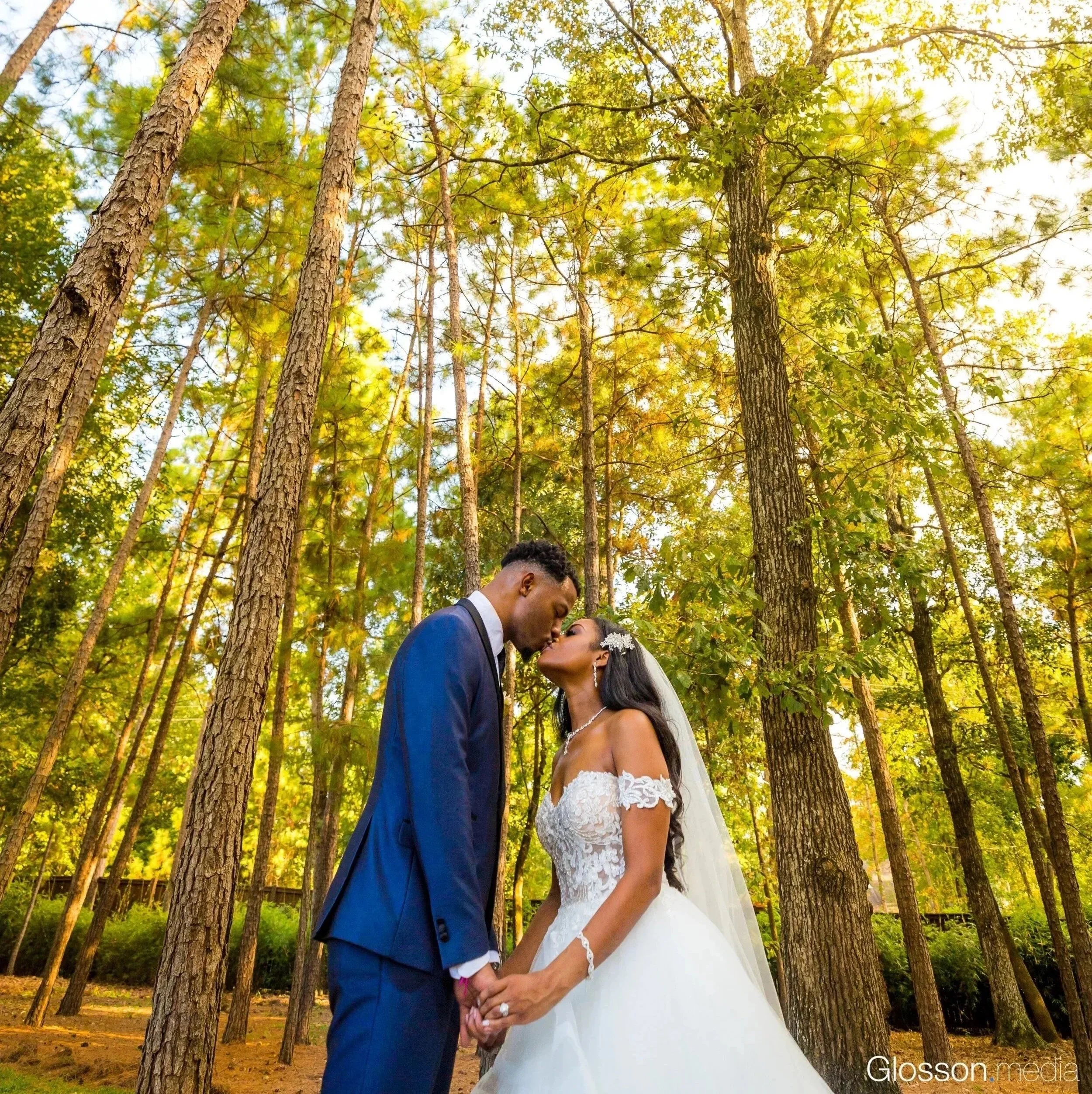 A bride and groom holding hands and leaning in for a kiss in a forest clearing with tall trees and sunlight filtering through.