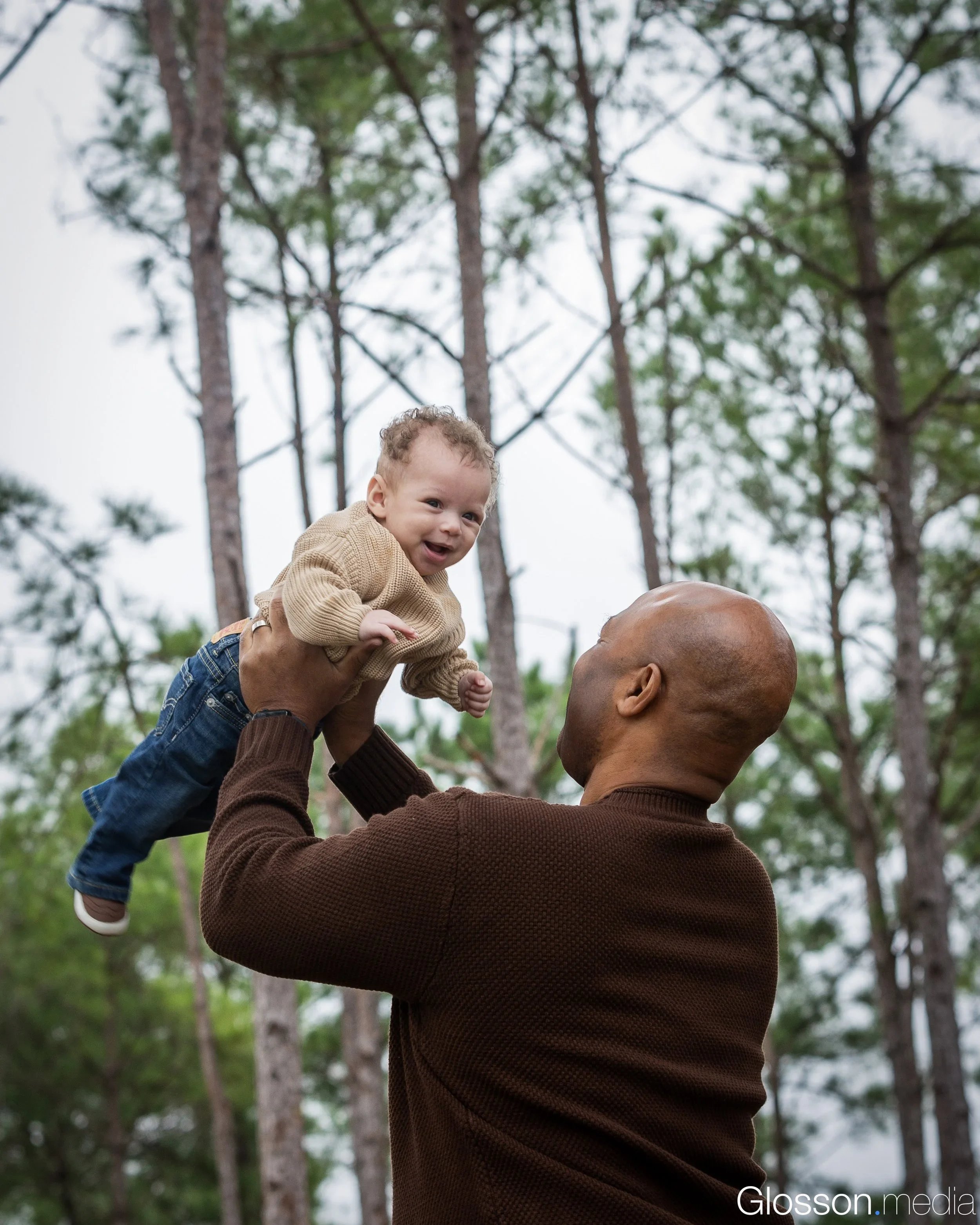 An adult man lifting a happy baby boy in the air outdoors, tall trees in the background.