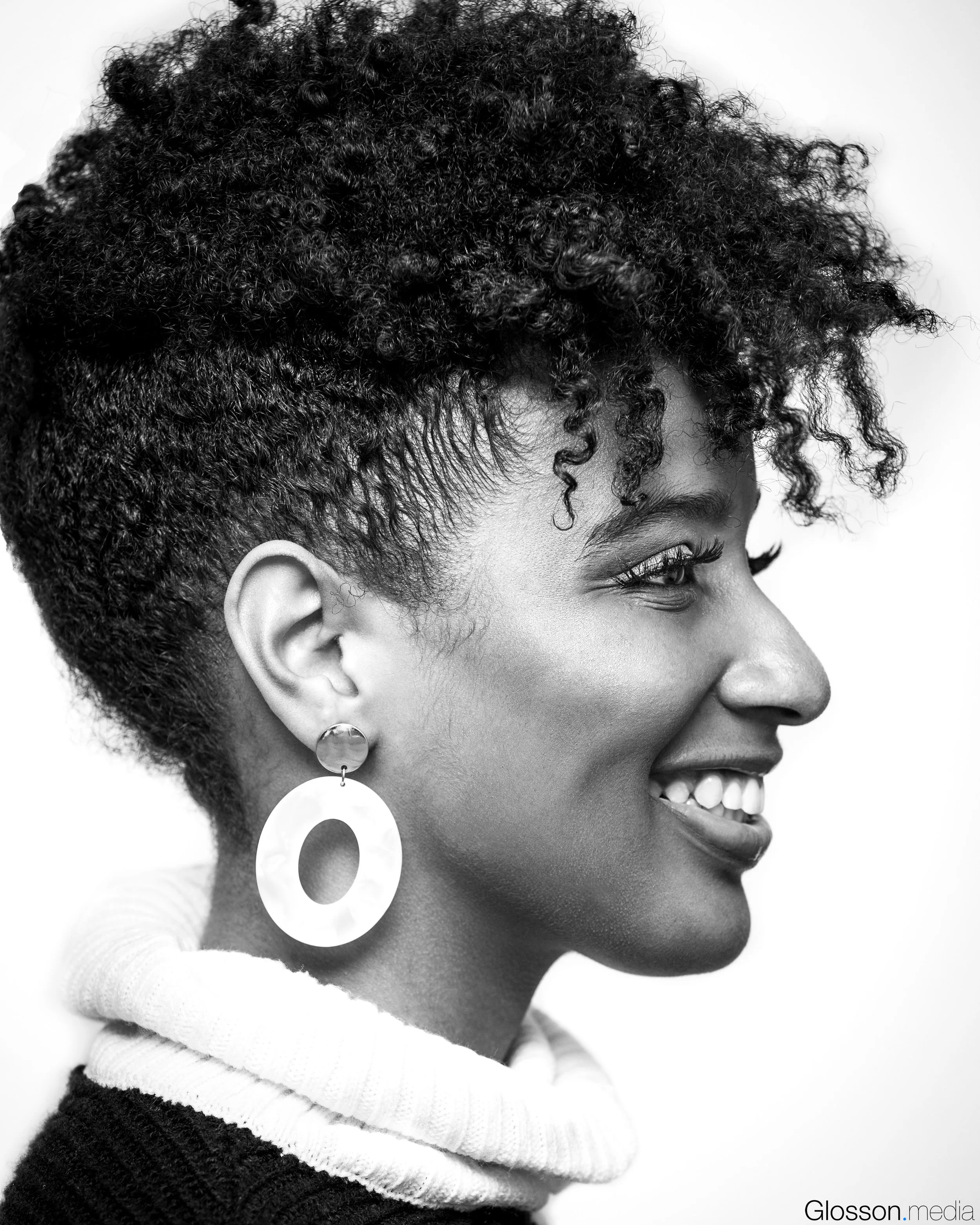 A black and white side profile of a smiling woman with short, curly hair, wearing large circular earrings and a turtleneck sweater.