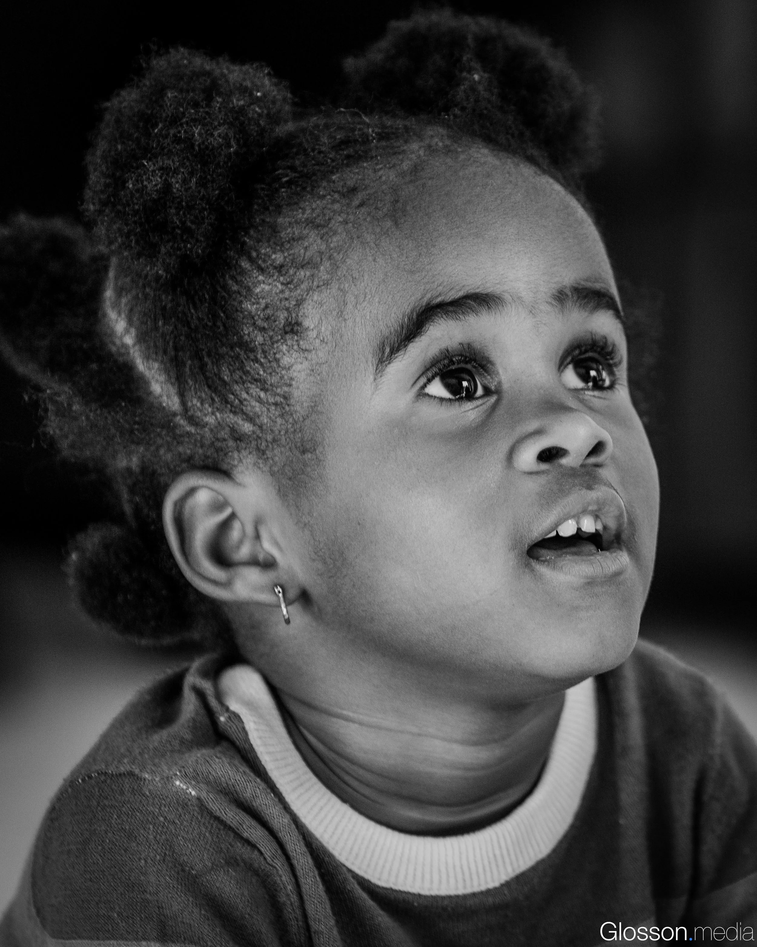 A close-up black and white photograph of a young girl with curly hair, looking upwards with wide eyes and a slight smile. She has earrings and is wearing a sweater over a collared shirt.