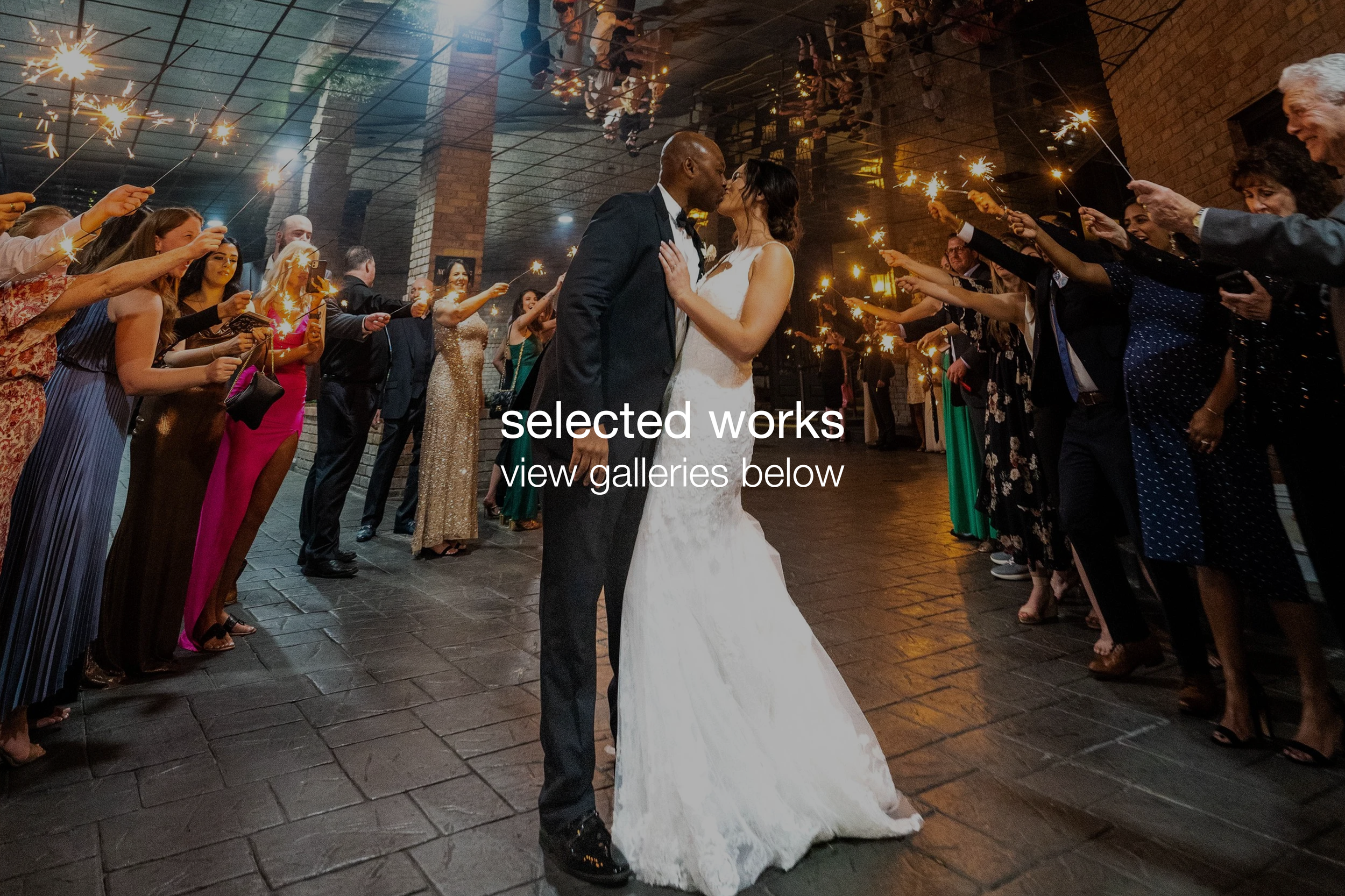 A newlywed couple kisses while holding sparklers during their wedding reception. Guests surround them, also holding sparklers, in a dimly lit indoor venue with a brick wall background.