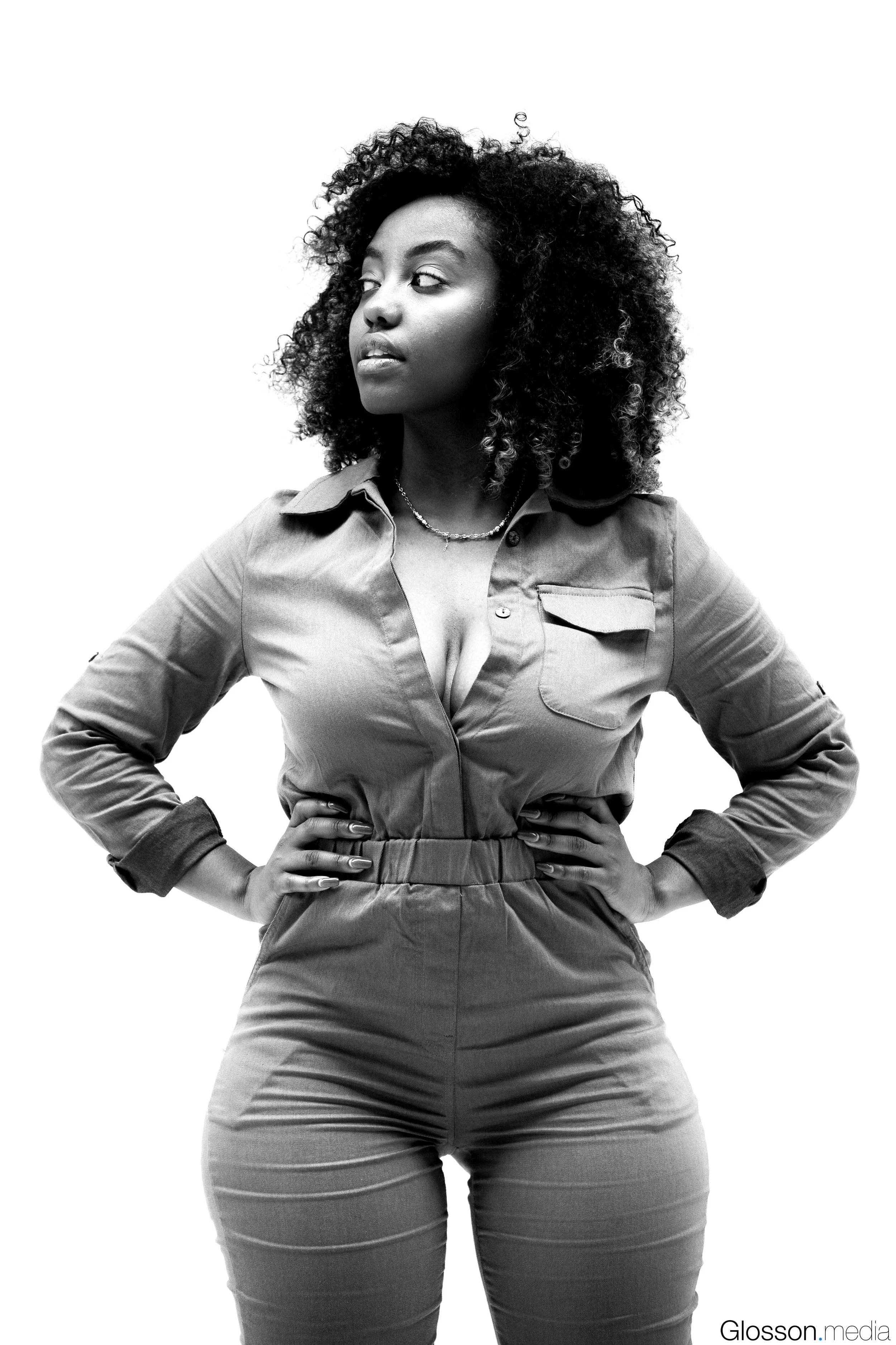 Black and white photo of a confident woman with curly hair, wearing a buttoned jumpsuit, with hands on hips, gazing to the side.