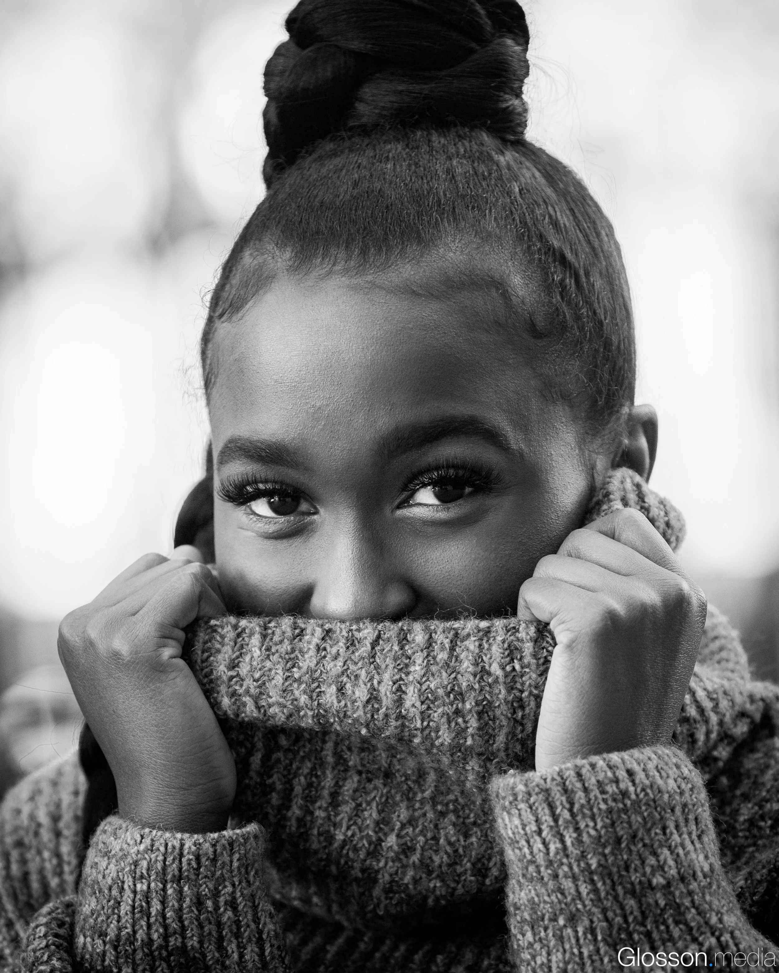 A close-up black-and-white photo of a woman with dark skin, short hair, and expressive eyes. She is wearing a thick knitted sweater and has her hands resting near her face, partially covering her cheeks.