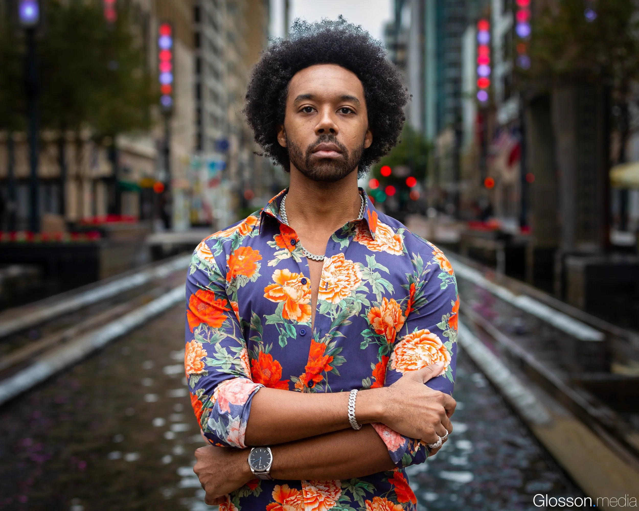 A man with dark skin and an Afro hairstyle wearing a colorful floral shirt and silver jewelry, crossing his arms, standing on train tracks in an urban city with buildings and neon lights in the background.