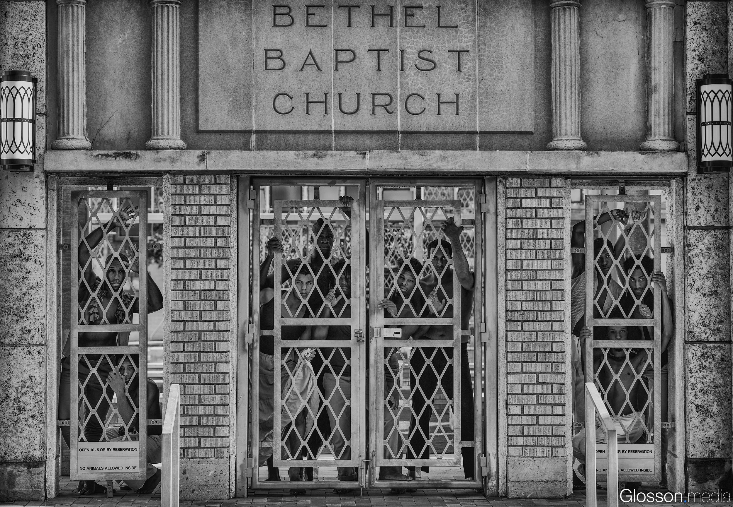 A group of people, including children, are gathered behind metal gates outside Bethel Baptist Church. The church's sign is visible above the entrance, and the building's architecture features columns and brickwork. Some individuals are looking throug