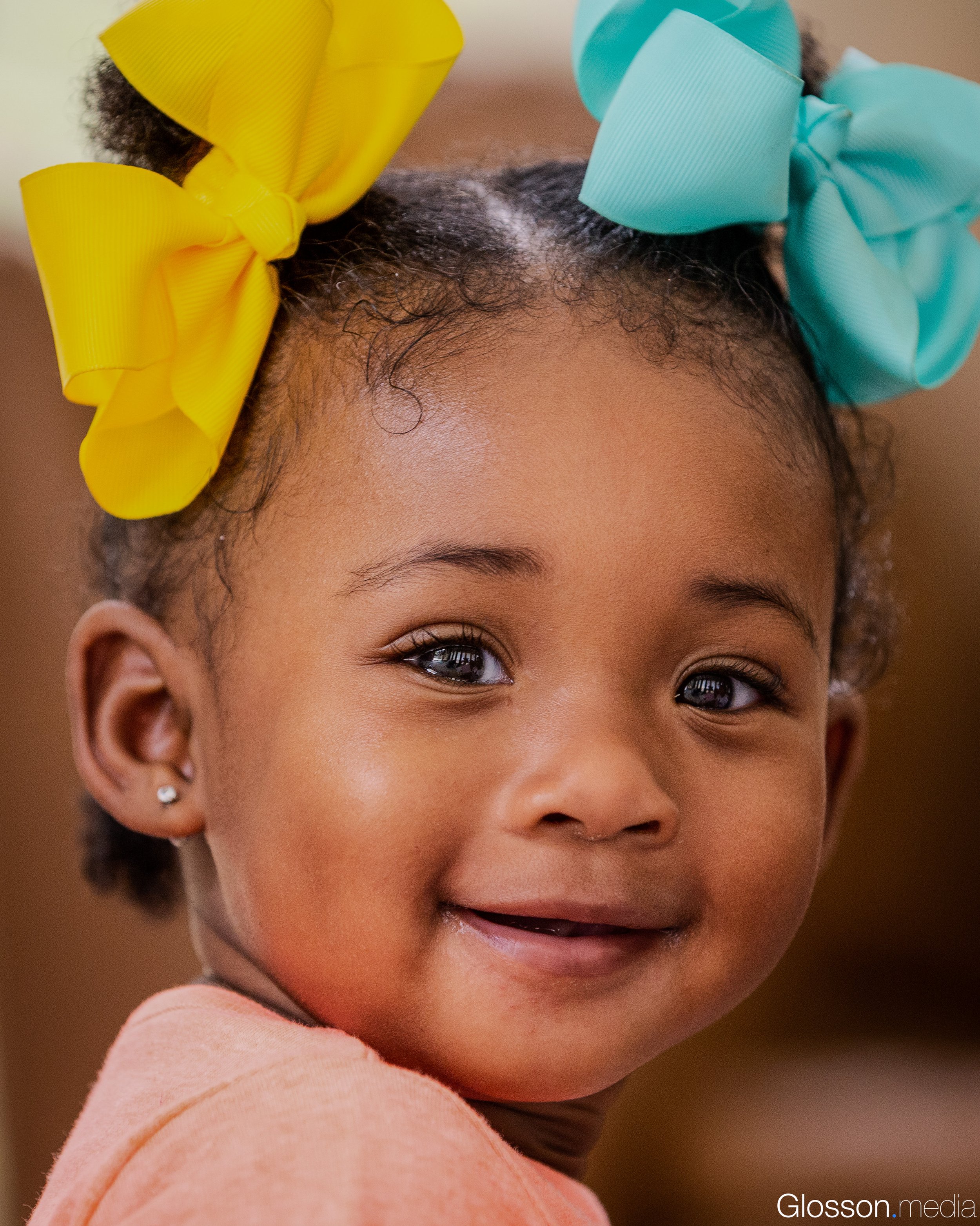 Close-up of a young girl smiling with curly hair tied with two large yellow and turquoise bows, wearing a peach-colored shirt.