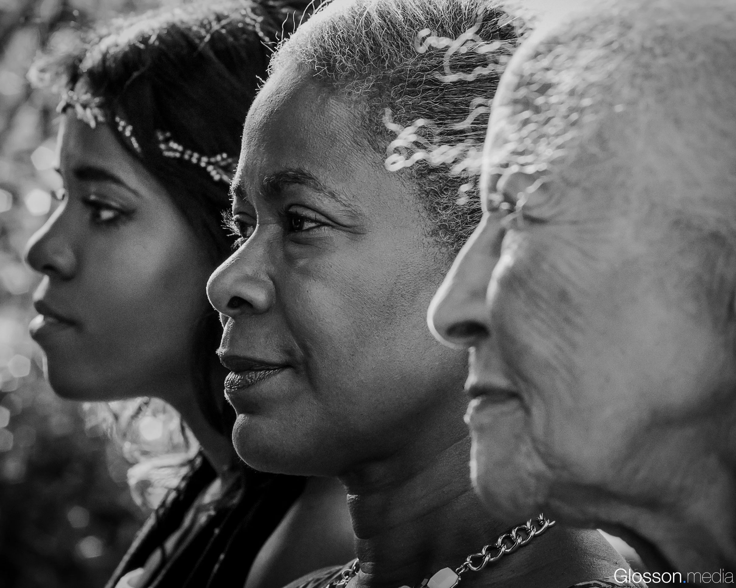 Close-up black and white photograph of three women’s faces in profile, with one woman in the foreground wearing a chain necklace and a keyed bracelet, and the background woman's face partially visible.