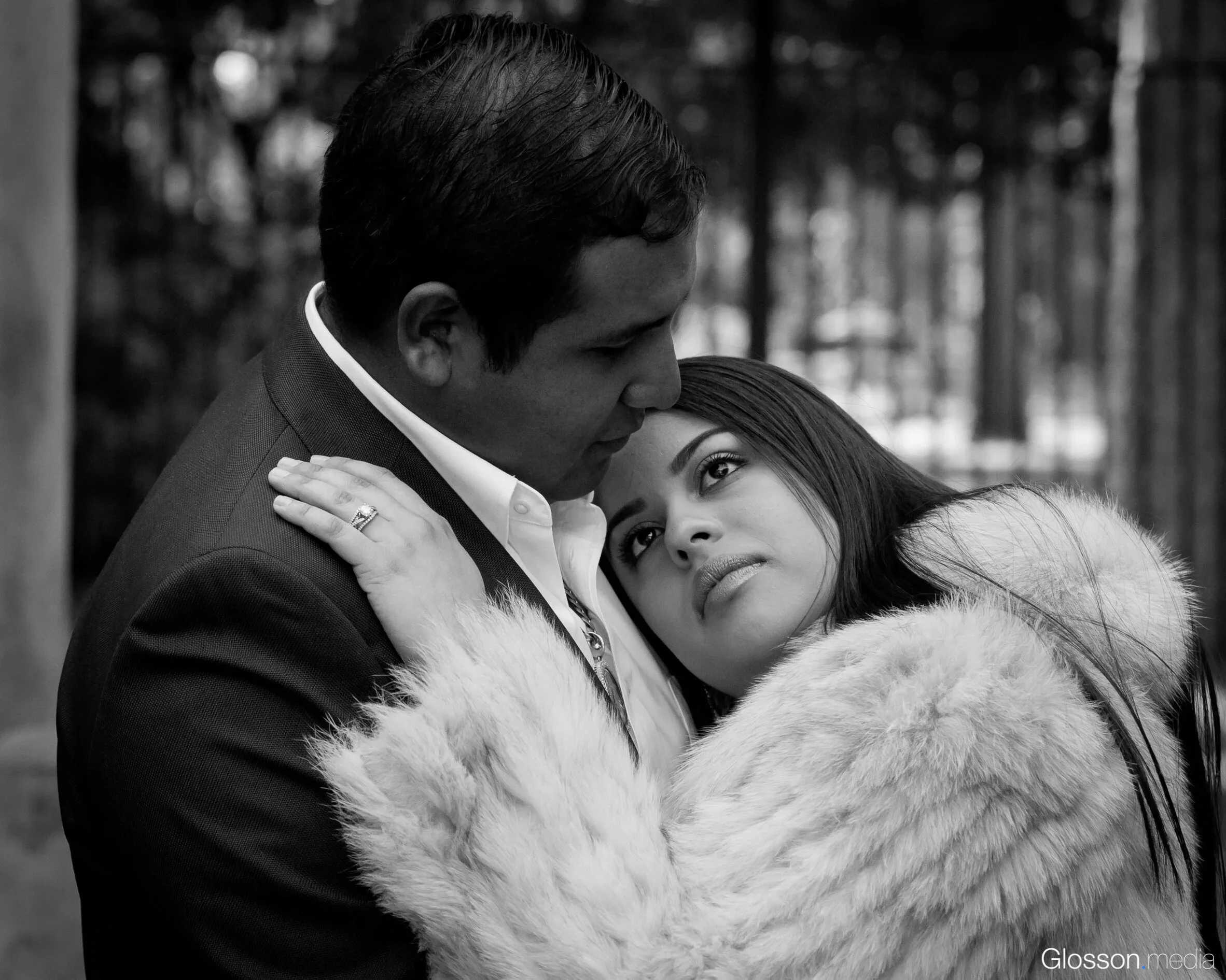A man and woman embrace outdoors, with the woman gazing into the man's face. The woman is wearing a fur coat, and the man is dressed in a suit.