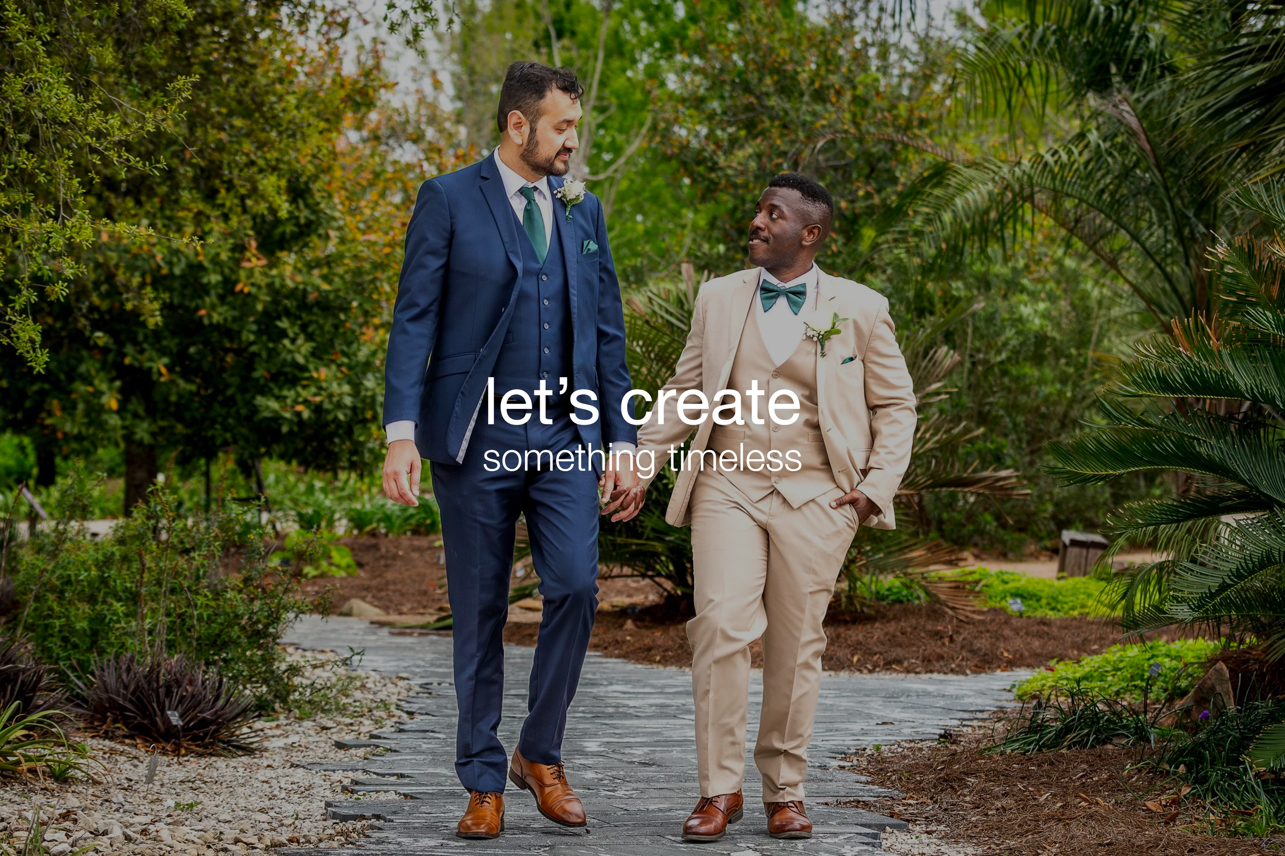 Two men in suits walking hand in hand in a garden, one in a navy suit and the other in a beige suit, holding hands and smiling at each other.