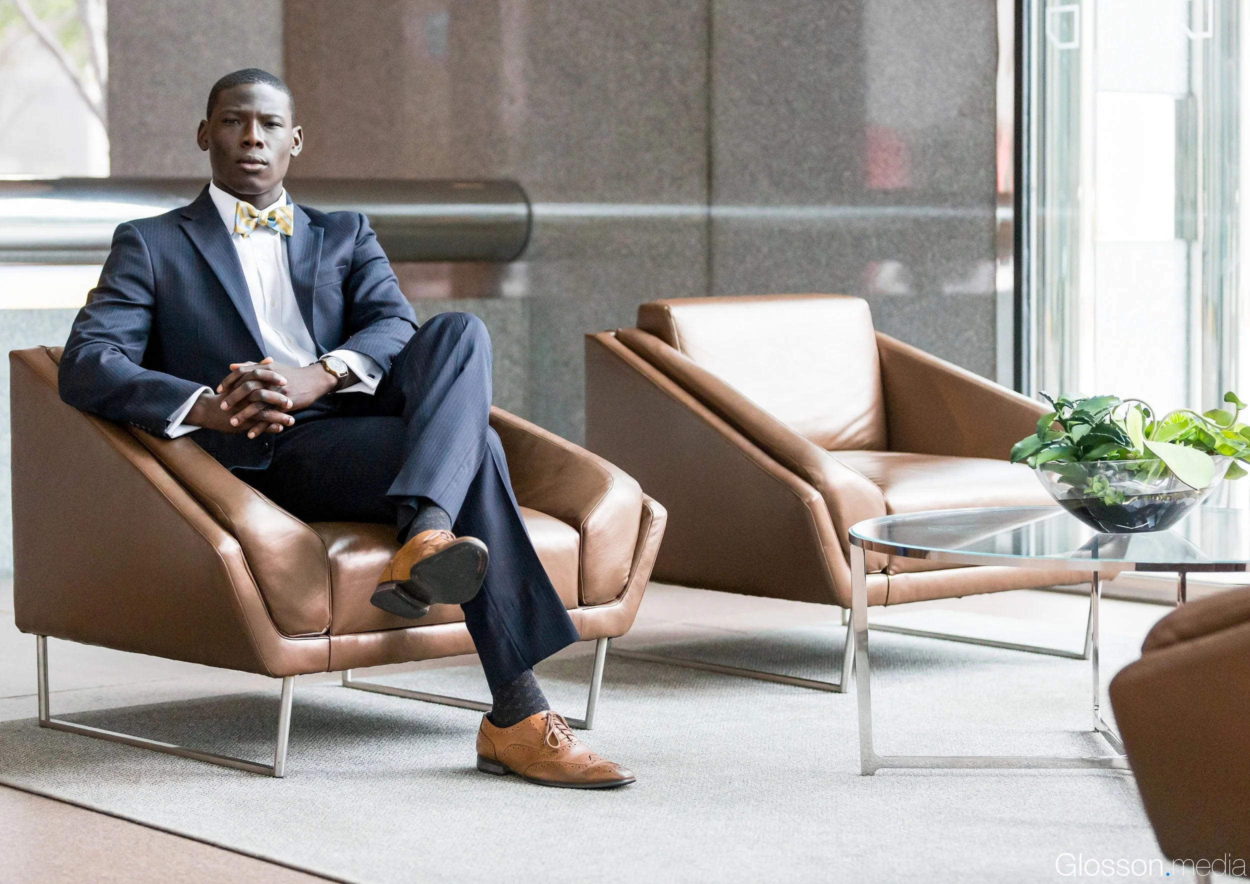 A young man in a formal suit with a yellow tie sits confidently in a modern lobby area, with brown leather chairs and a glass table with a decorative plant, by a large window with natural light.