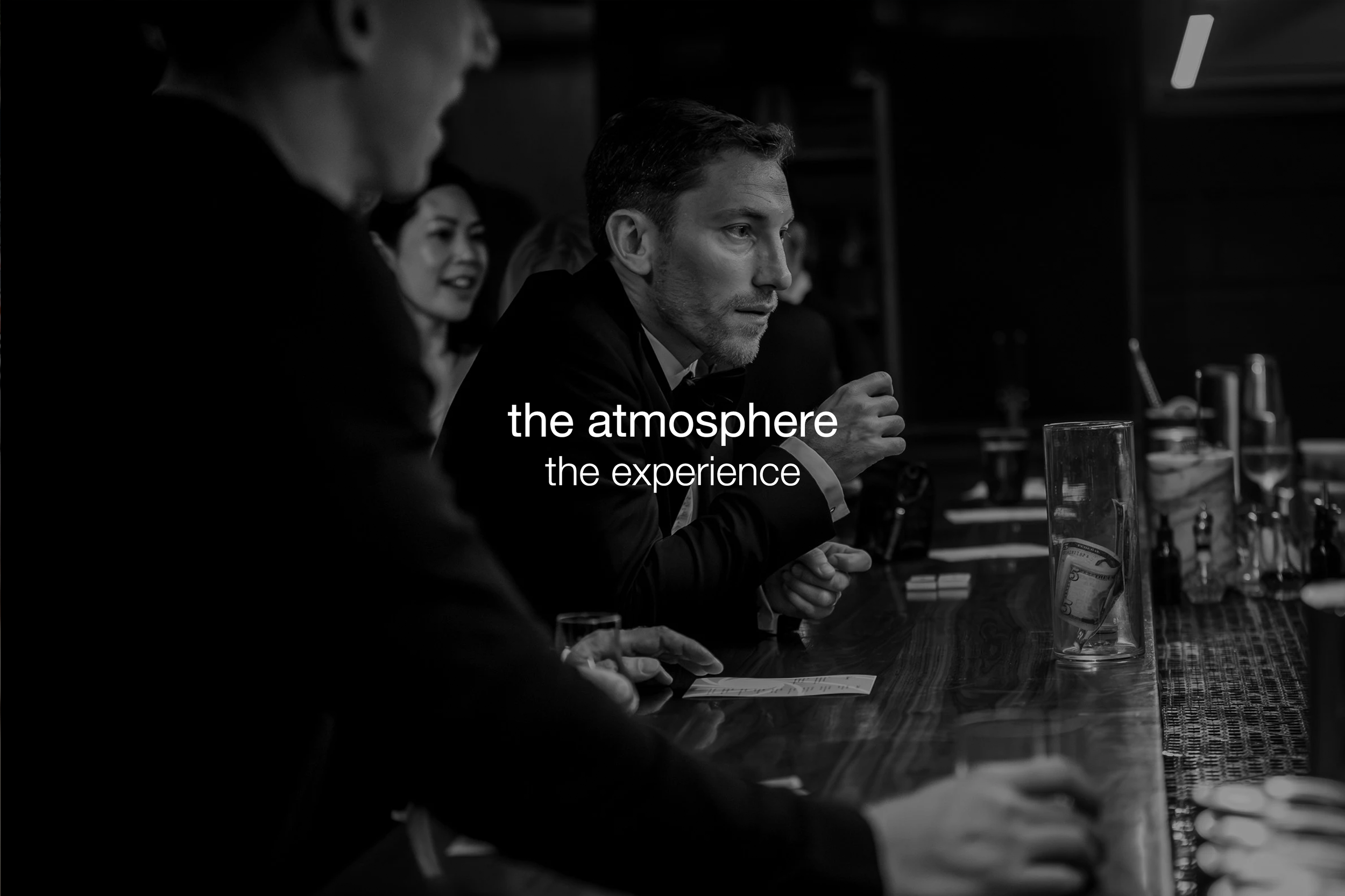 A group of people dressed in formal attire sitting at a long bar counter, engaged in conversation, with drinks and glasses in front of them, in a dimly lit setting.