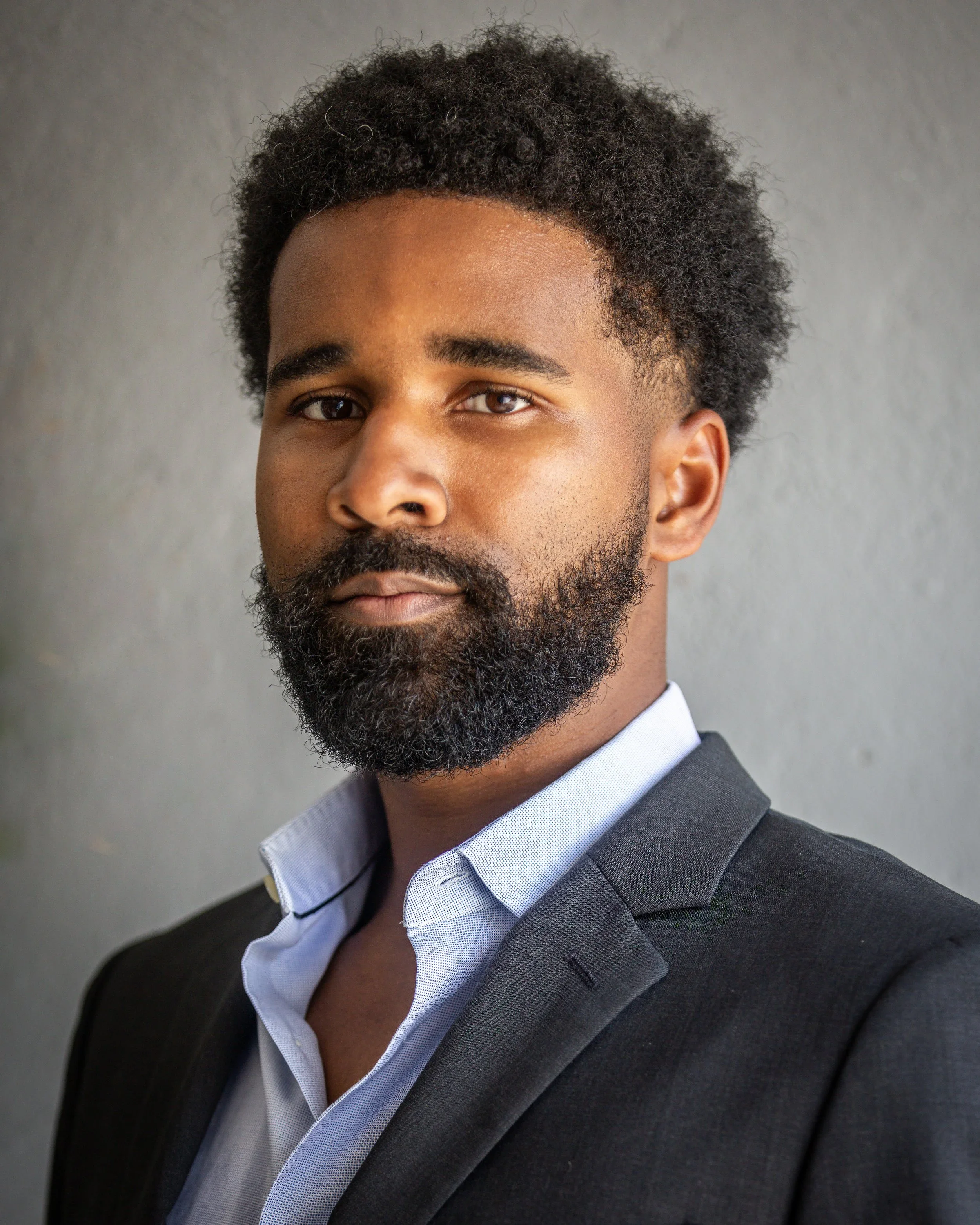 Close-up photo of a man with dark curly hair, thick beard, wearing a dark suit and light blue shirt with a collar, against a gray background.