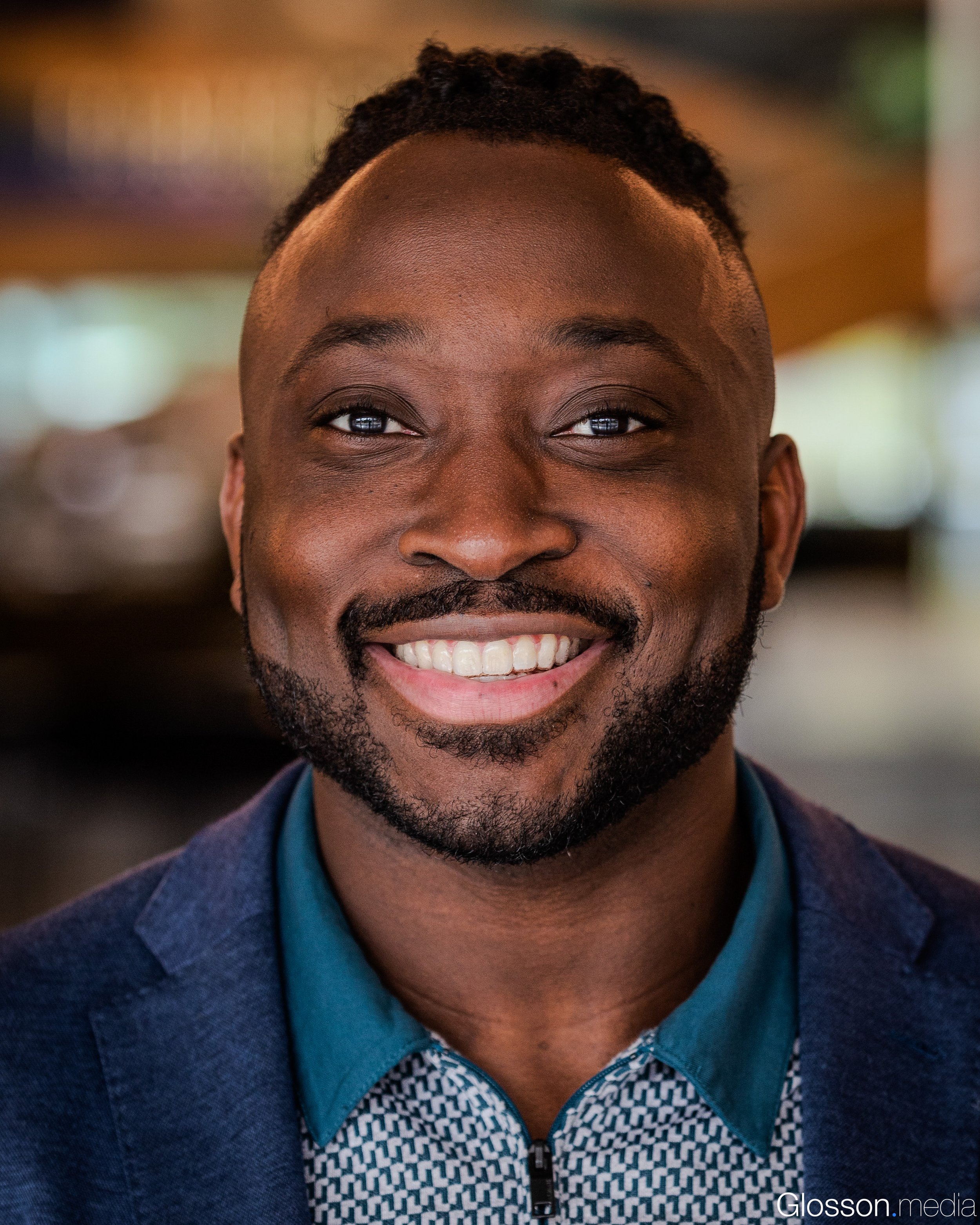 A young man with dark skin, short curly hair, and a beard, smiling with teeth showing. Wearing a blue shirt and a dark blazer. The background is blurred with warm lighting.