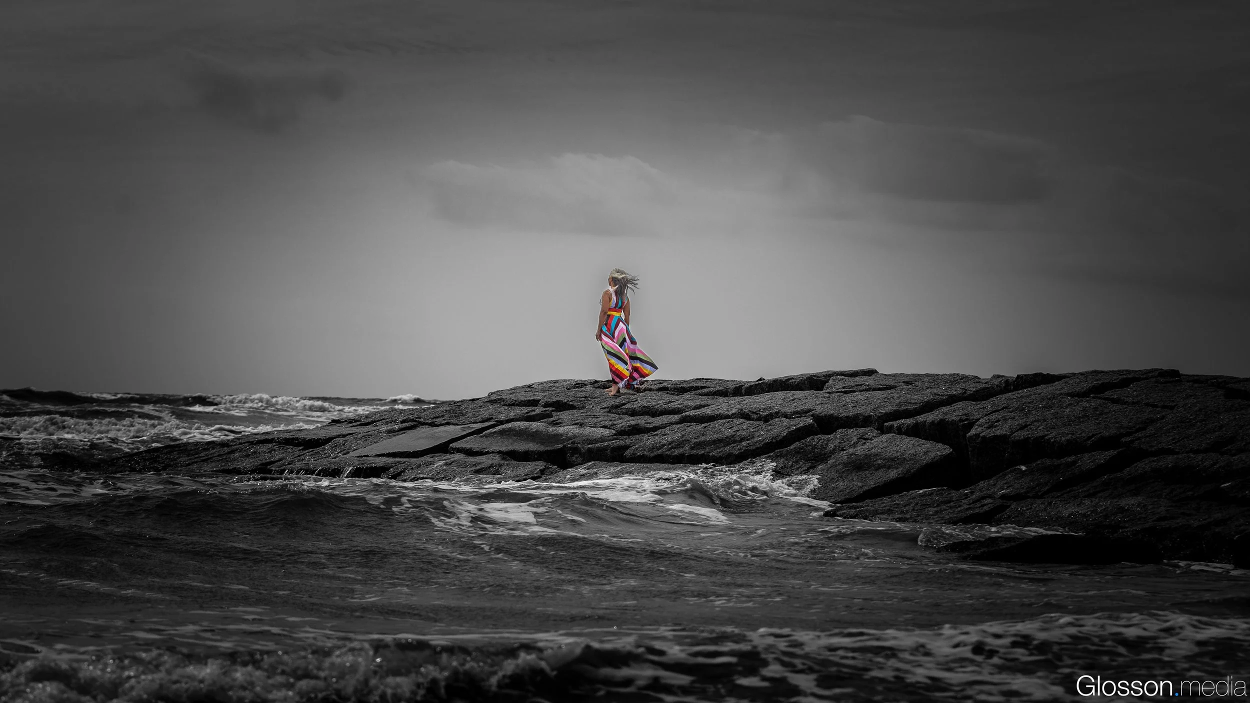 A woman in a colorful striped dress walking on rocks near the ocean on a cloudy day, with the image primarily in black and white except for the dress.