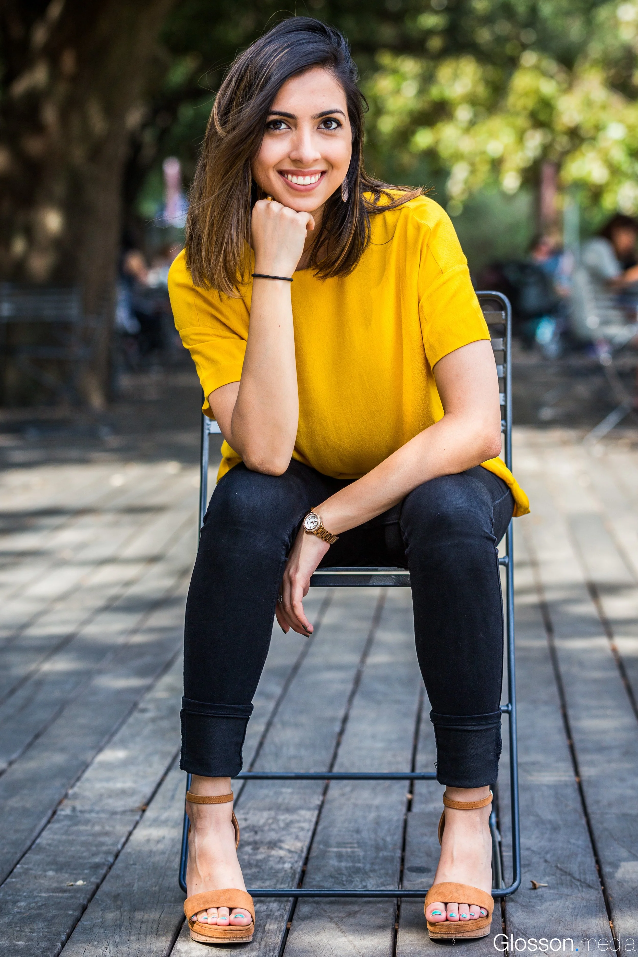Young woman sitting outdoors on a wooden deck, smiling with her chin resting on her hand, wearing a yellow top, black jeans, tan high-heeled sandals, with blurred trees and people in the background.