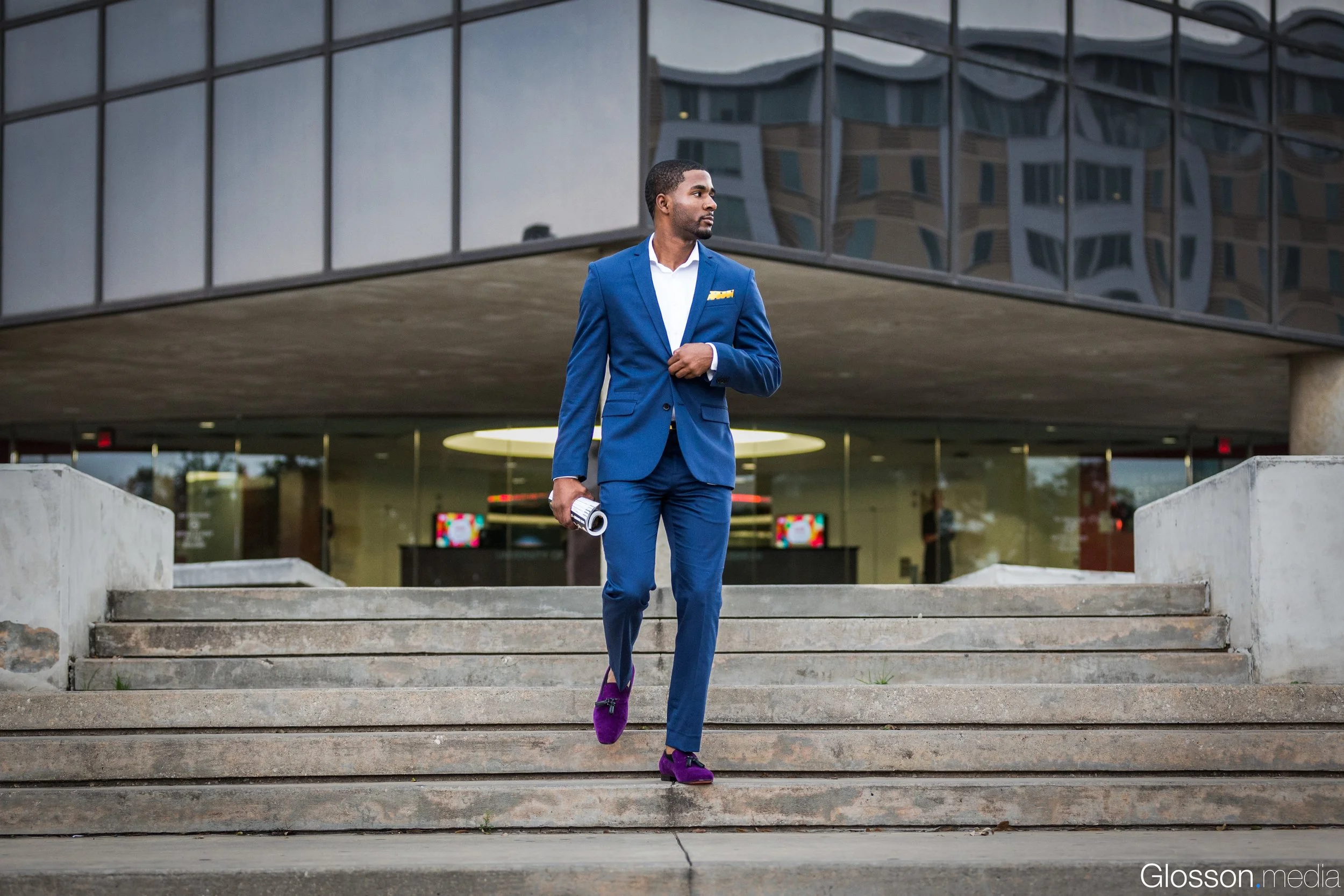 A man in a sharp blue suit with a yellow pocket square walks down concrete stairs outside a modern building with reflective glass windows. He is holding a rolled-up document in his right hand and wearing purple shoes.