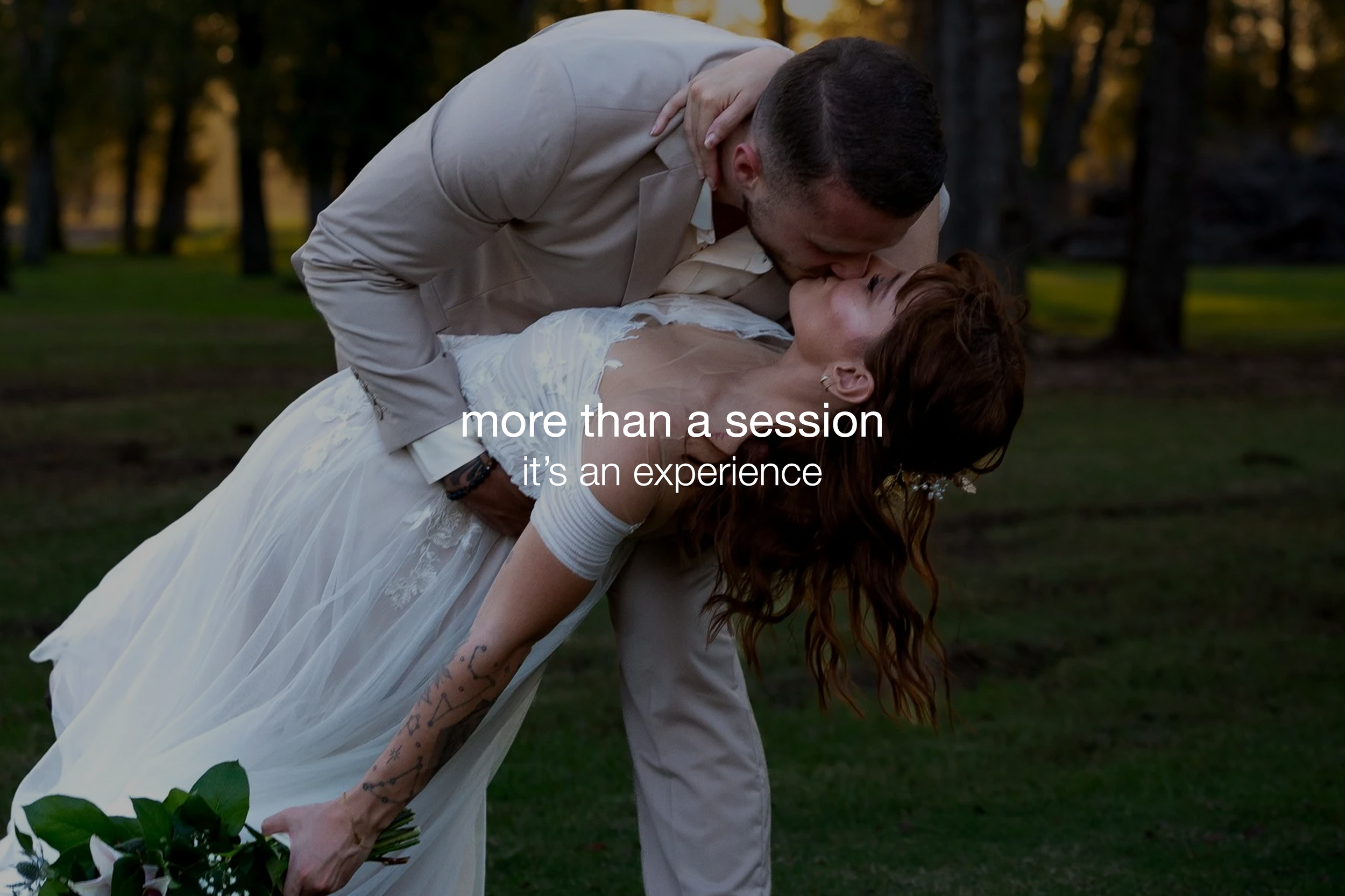 A couple dressed in wedding attire sharing a romantic kiss outdoors, with the man bending the woman backwards while she holds a bouquet of flowers.