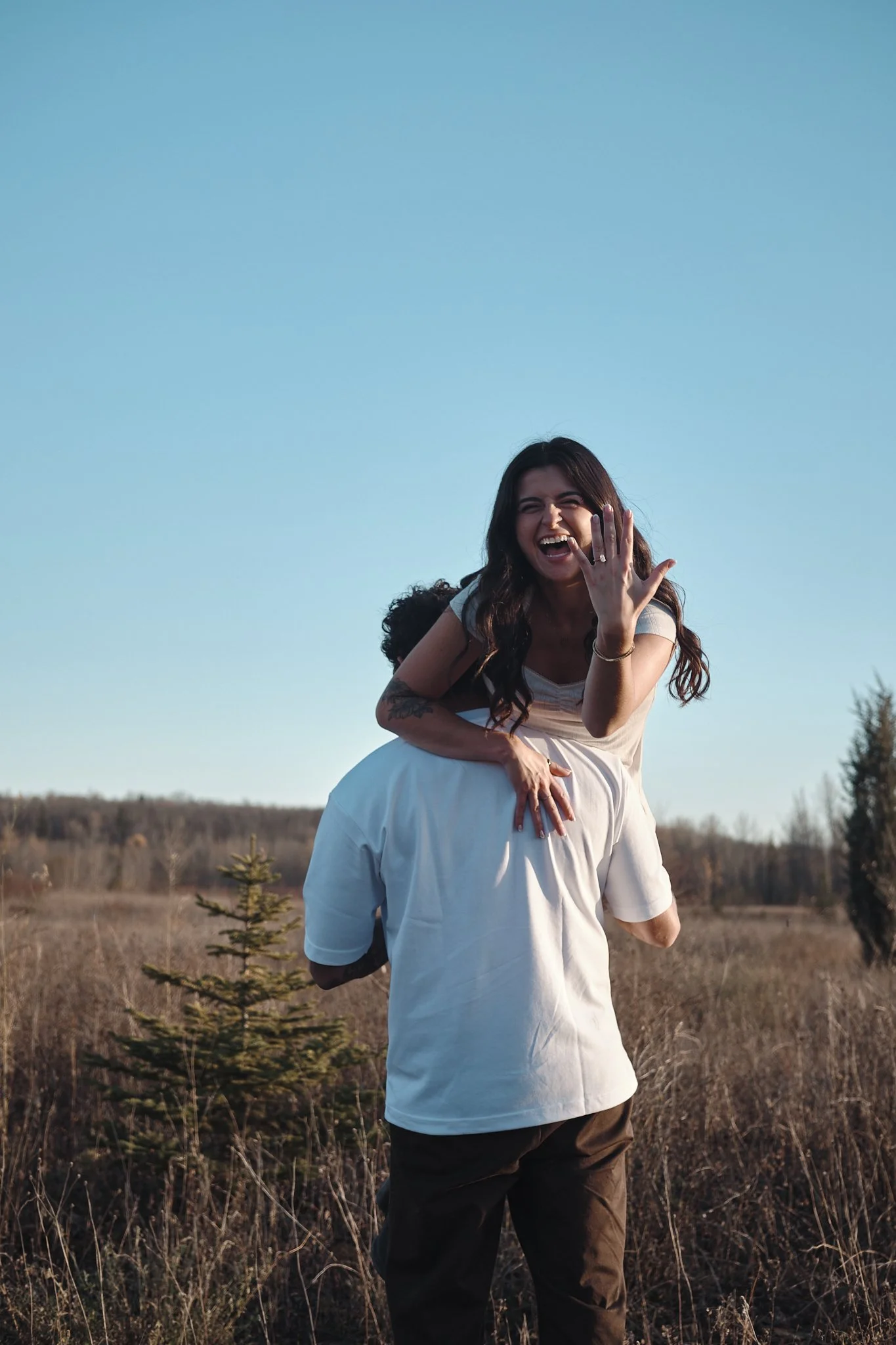 A woman in a white dress is being carried on a man's shoulders in a field outdoors, smiling and laughing, with a clear blue sky in the background. Taken in Edmonton, Alberta.
