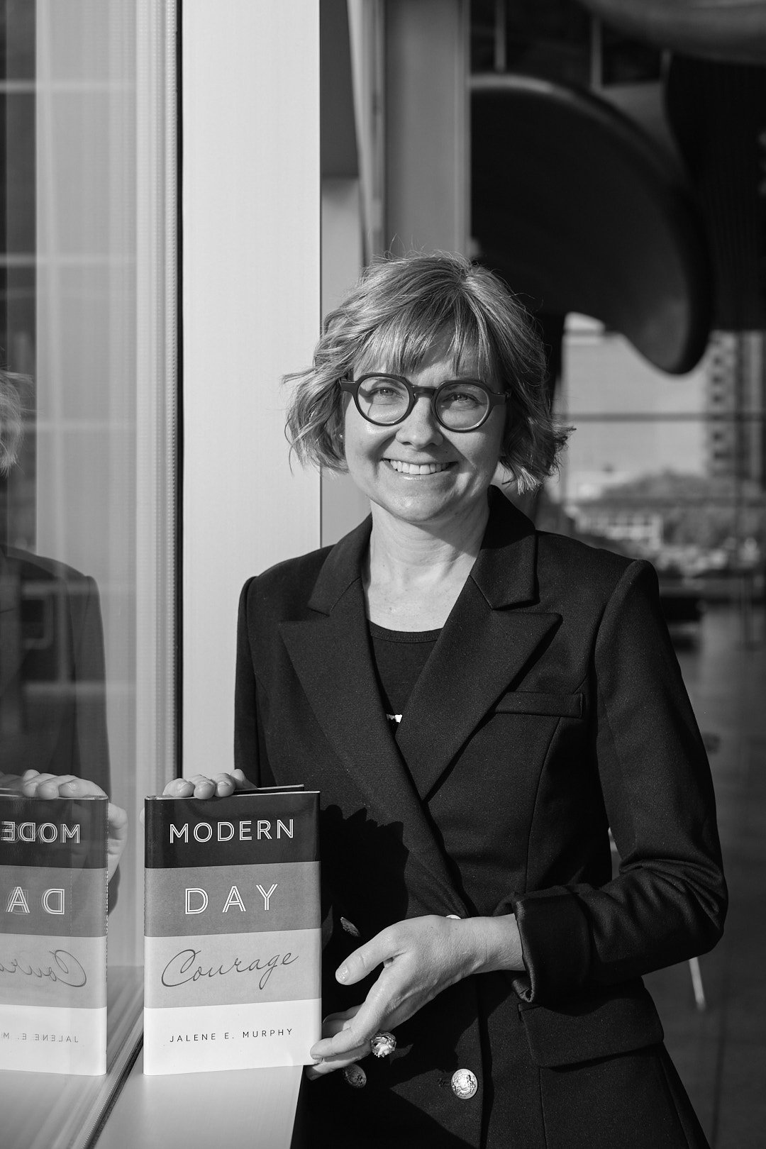 A woman with short blonde hair and glasses, wearing a dark blazer, standing outdoors next to a window with books titled 'Modern Day Courage' on display. She is smiling and looking to her left. Taken in Edmonton, Alberta.