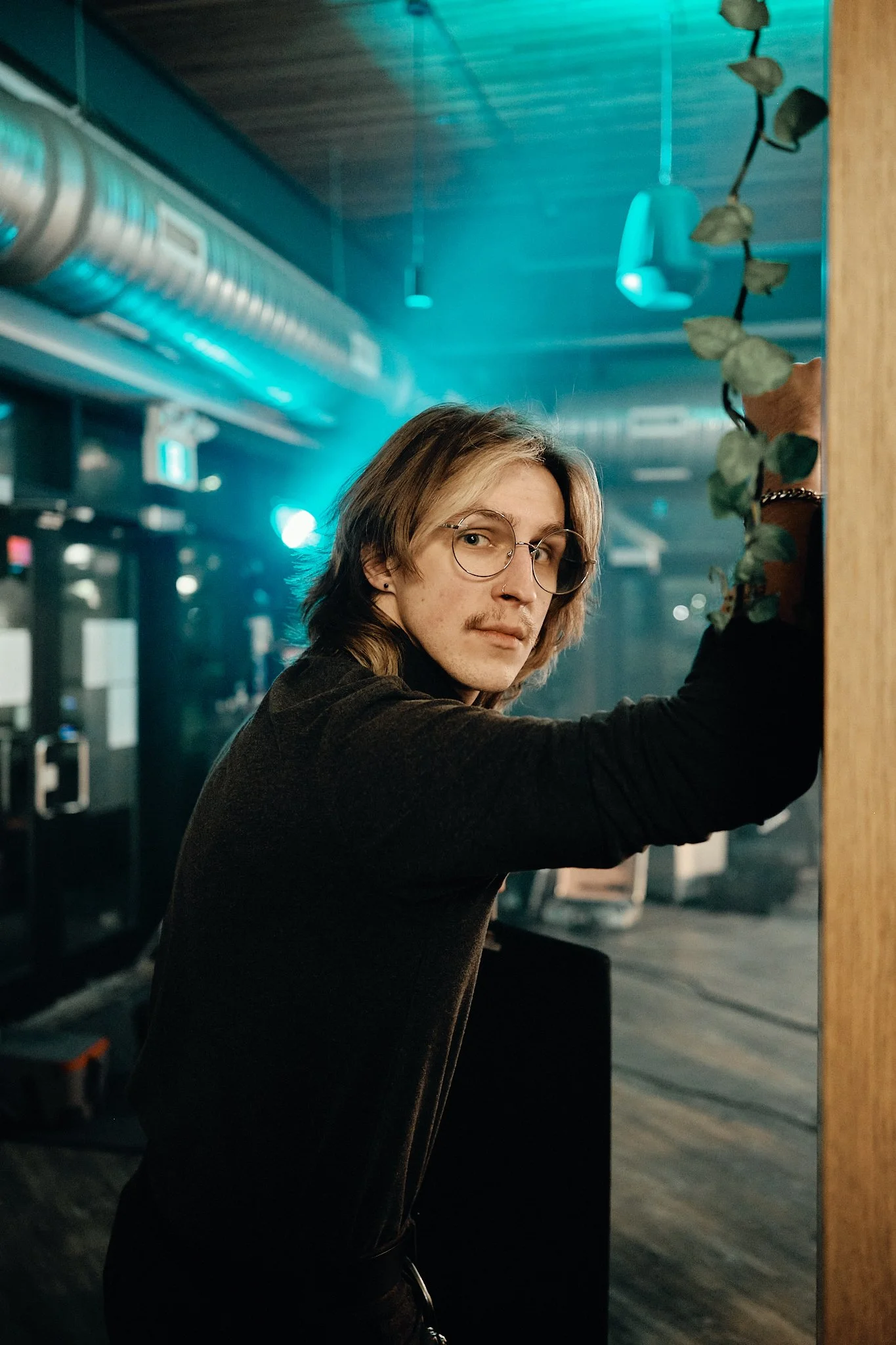 A young man with shoulder-length blonde hair, glasses, and a black long-sleeve shirt, standing indoors near a mirror with a plant in front of him, in a setting with blue lighting and visible industrial-style ducts on the ceiling. Taken in Edmonton, A
