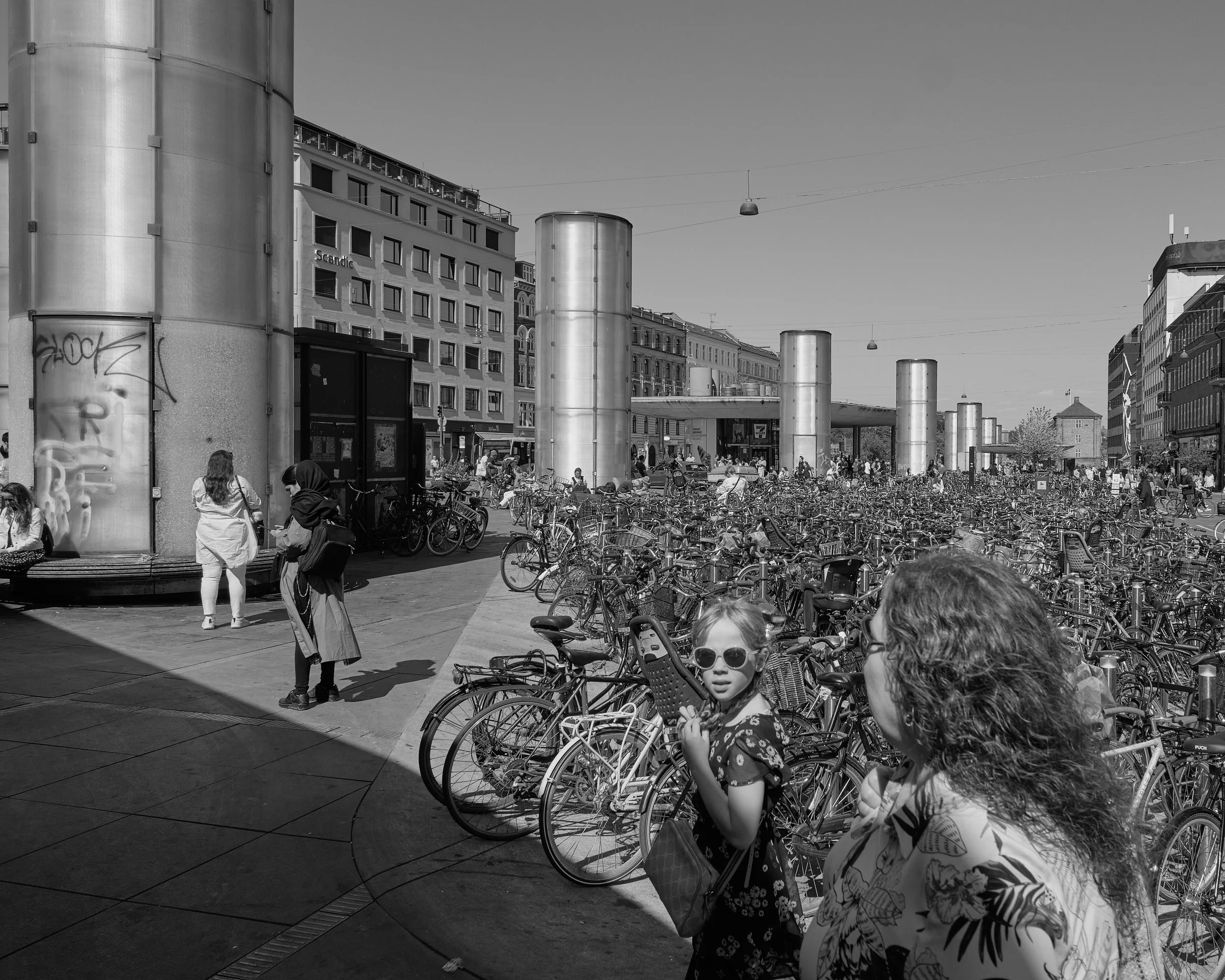 A city square in Copenhagen, Denmark, filled with bicycles, with pedestrians walking around. Modern buildings and large metallic columns are visible in the background, along with a few children and adults.