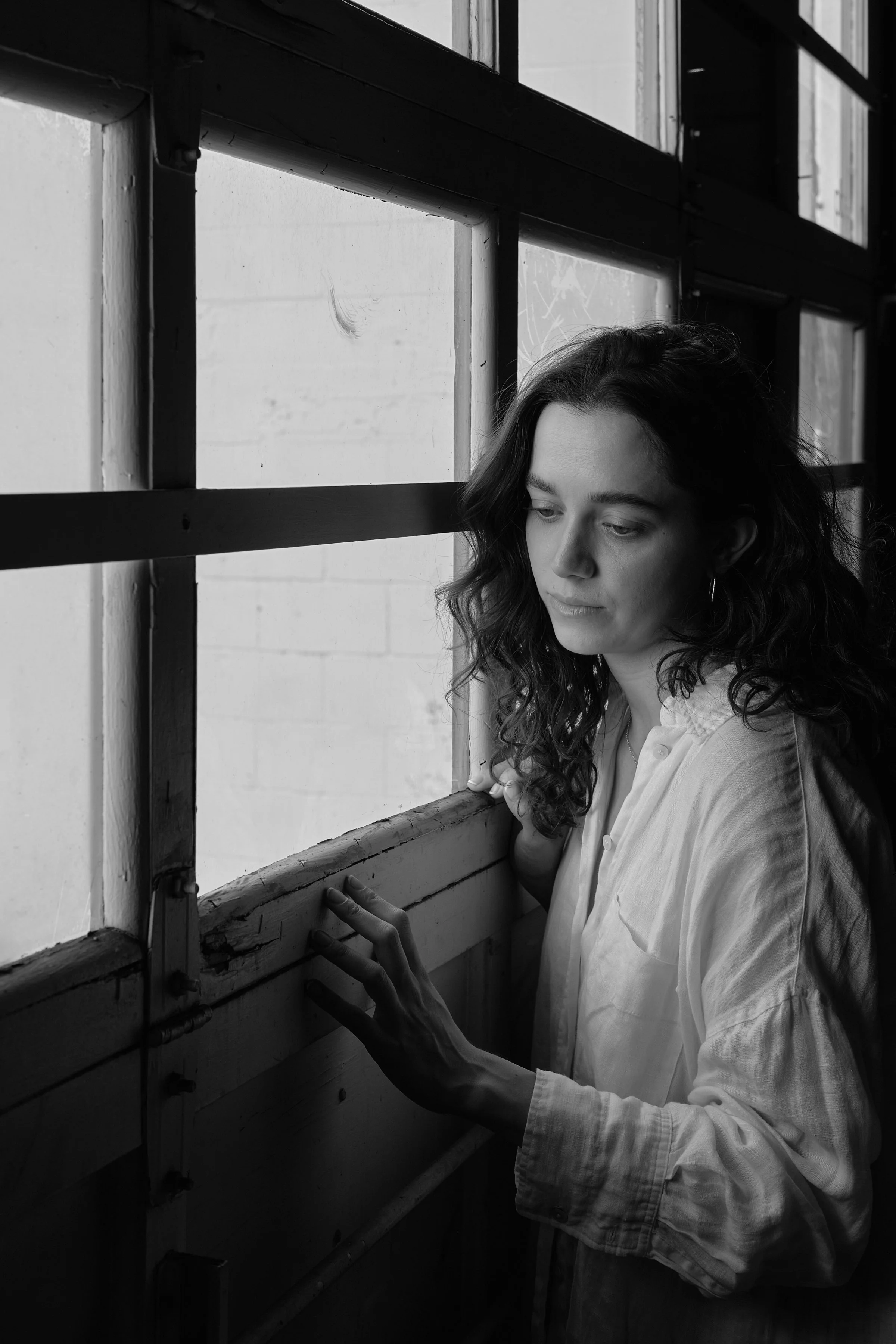 A woman with wavy hair wearing a light-colored shirt, standing by a window, looking down and touching the window frame with her right hand.