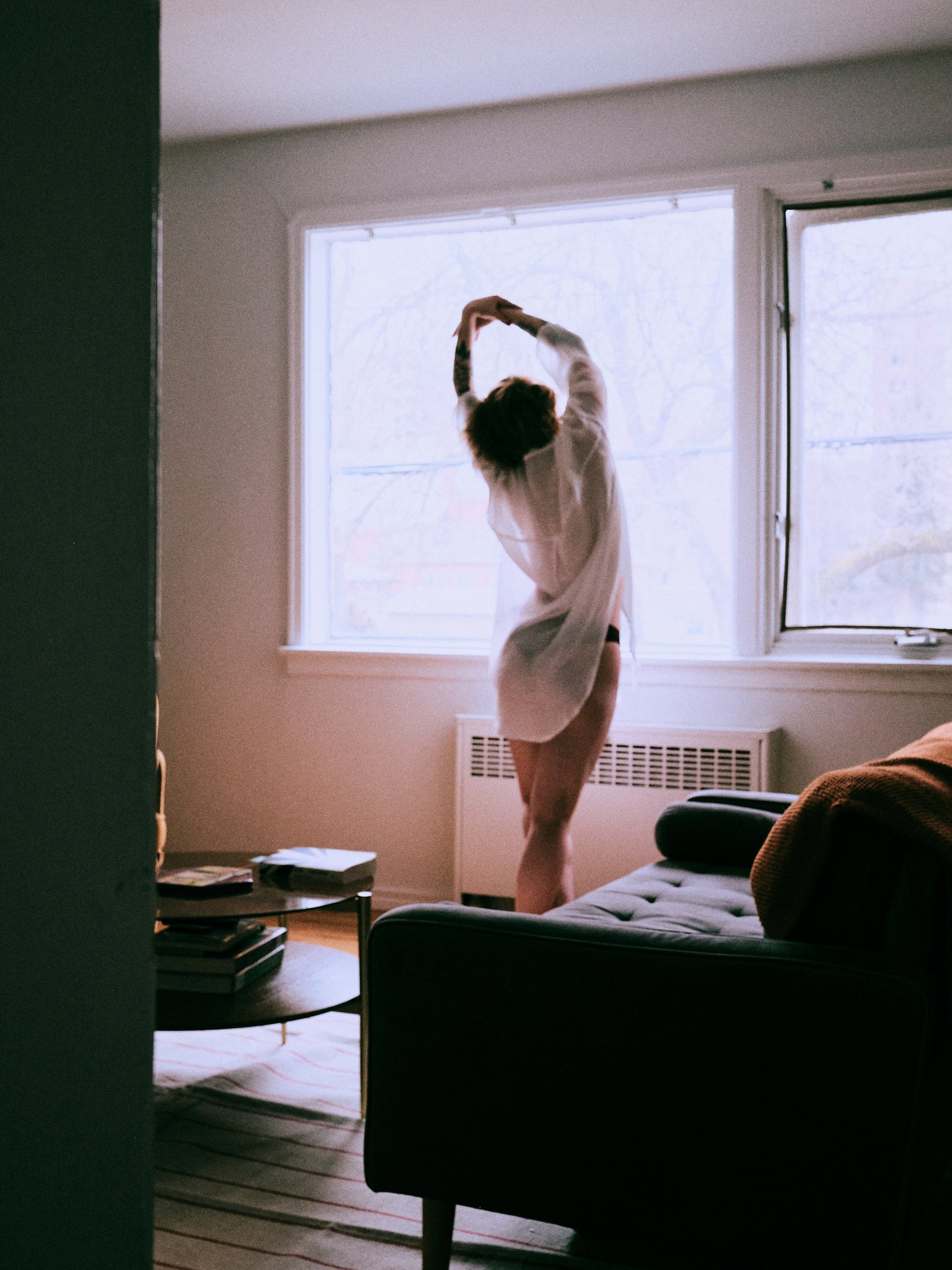 A person standing in a living room by a large window, stretching or dancing with arms raised above their head. The room has a radio and some books on a table, with a black couch and a bed visible.