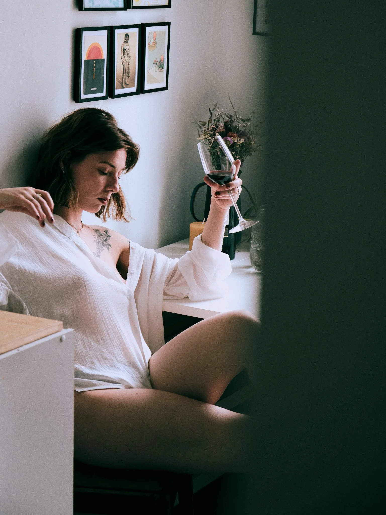 A woman with short brown hair and tattoos on her arm, wearing a white shirt, sitting in a room, holding a glass of red wine, with a framed art collection and a flower arrangement behind her.