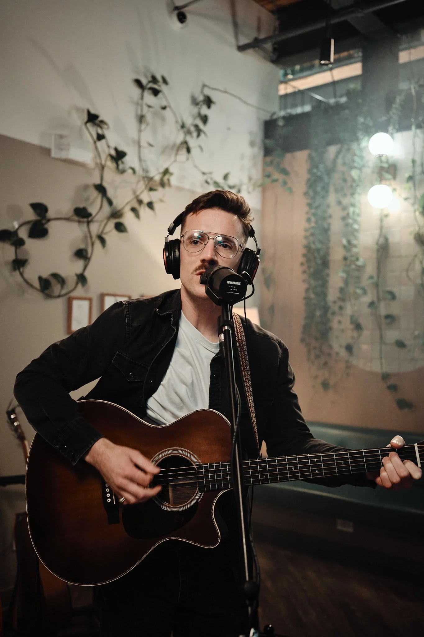 A man with short hair, glasses, and a mustache is singing into a microphone while playing an acoustic guitar in a cozy indoor setting with hanging lights and decorative plants. Taken in Edmonton, Alberta.