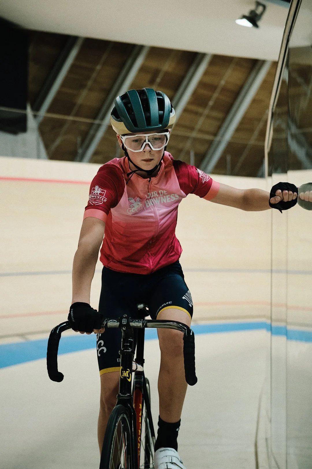 A young cyclist wearing a helmet, goggles, a pink and red cycling jersey, black shorts, and gloves, riding a bike indoors on a velodrome track. Taken in Edmonton, Alberta.