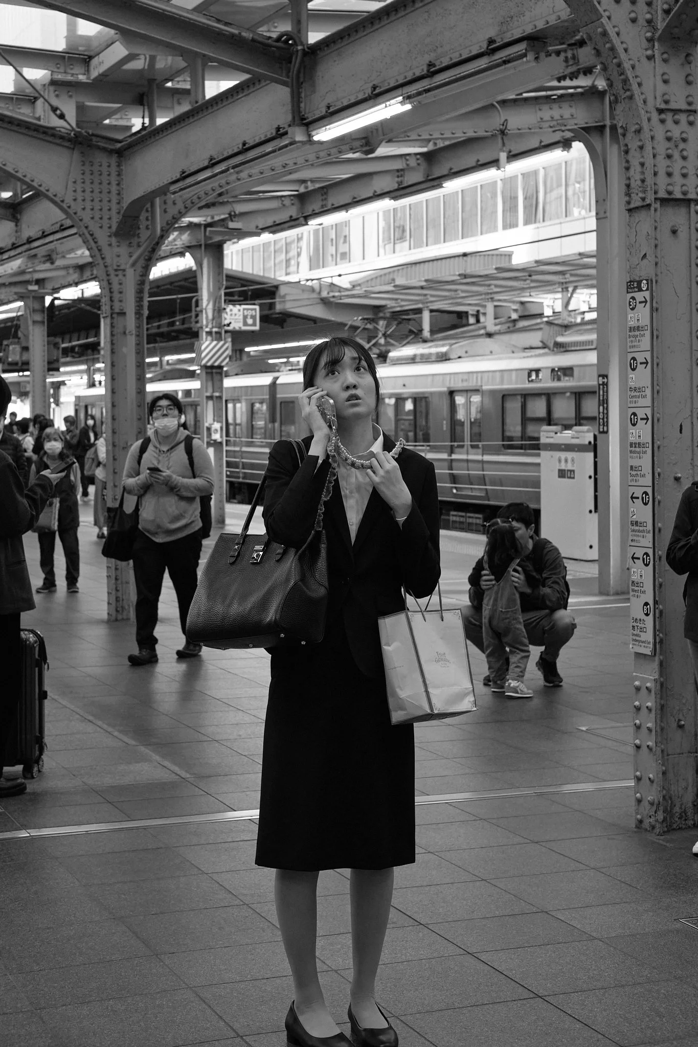 A woman in professional attire standing on a busy train platform in Japan, talking on her mobile phone, surrounded by other travelers, with a train in the background and steel infrastructure overhead. 