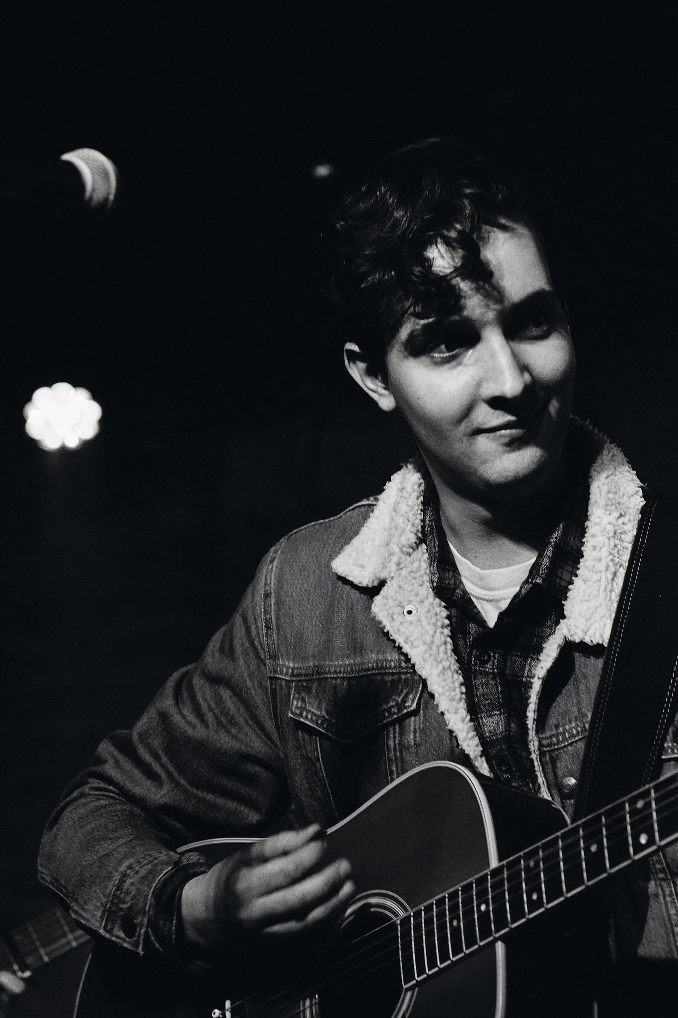 A young man with dark, curly hair playing an acoustic guitar. He is wearing a denim jacket with a shearling collar and a plaid shirt underneath. The background is dark with some blurred stage lights. Edmonton, Alberta