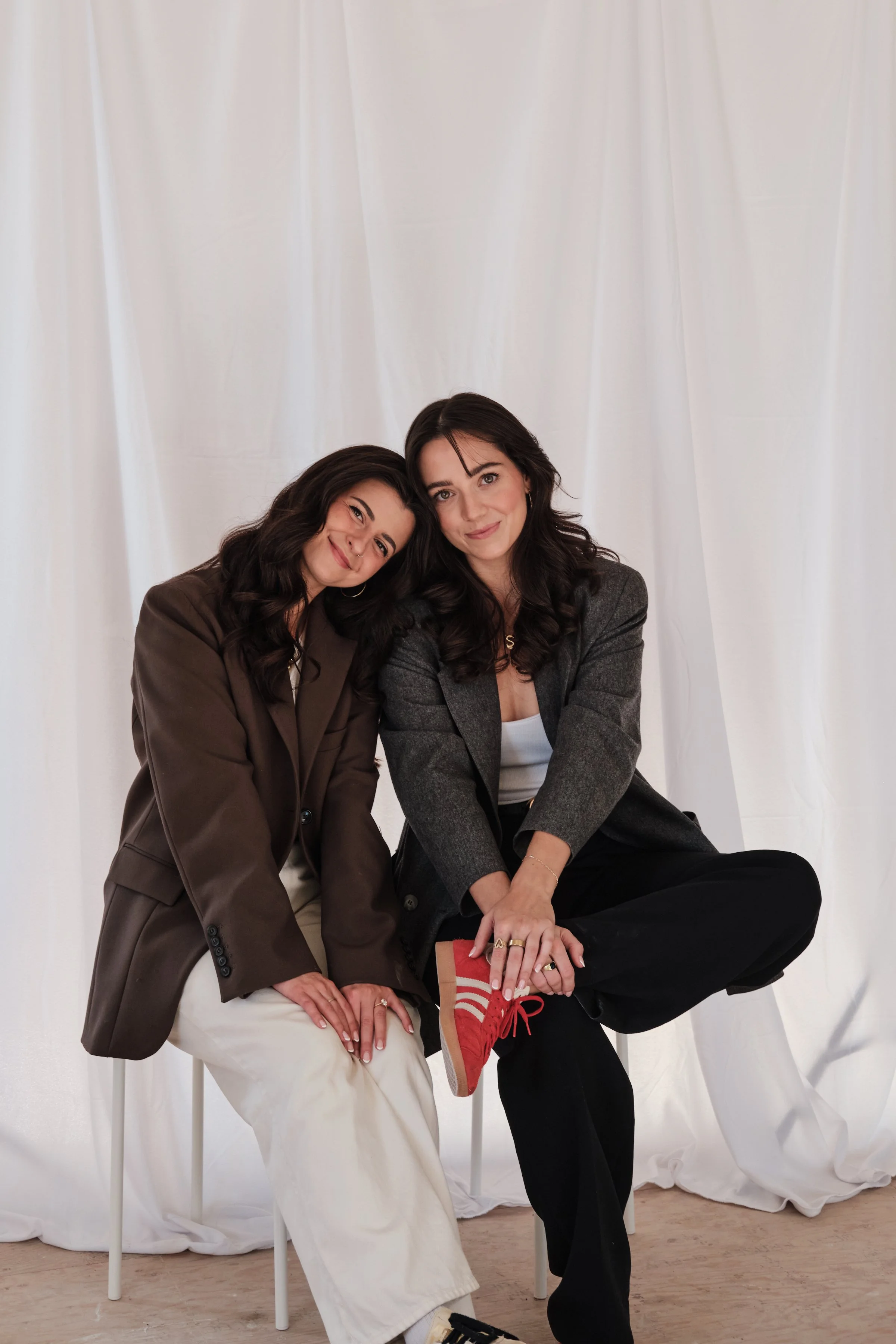 Two women sitting together on chairs in front of a white curtain backdrop, smiling at the camera. One woman wears a brown blazer and white pants, and the other wears a gray blazer, black pants, and red sneakers. Taken in Edmonton, Alberta.