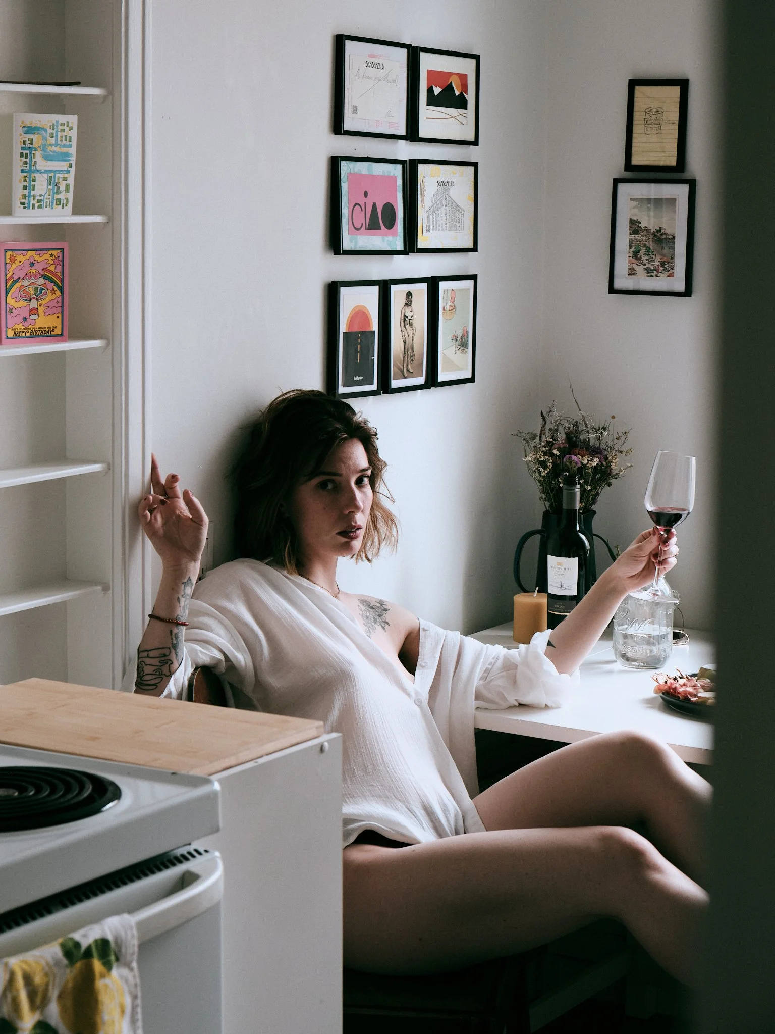 A woman with tattoos on her arm, sitting on a kitchen table, holding a glass of red wine, and looking at the camera, with a wall decorated with framed artwork behind her, a plant, a bottle of wine, and a candle on the table.