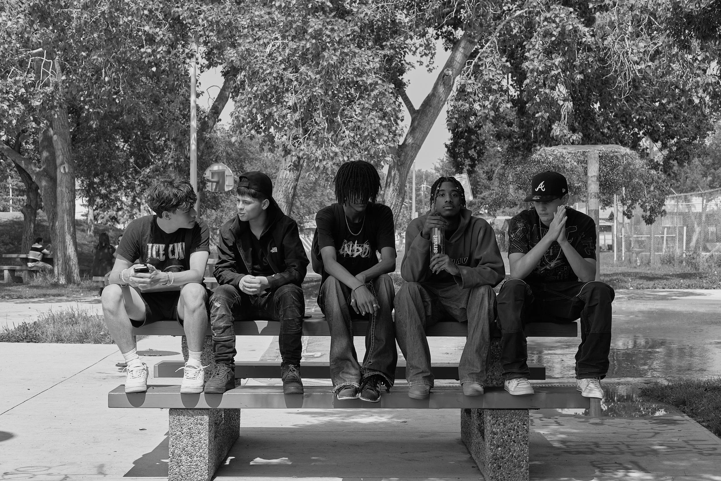 Five young men sitting on a bench outdoors, with trees and a fence in the background, some using their phones and one holding a water bottle.